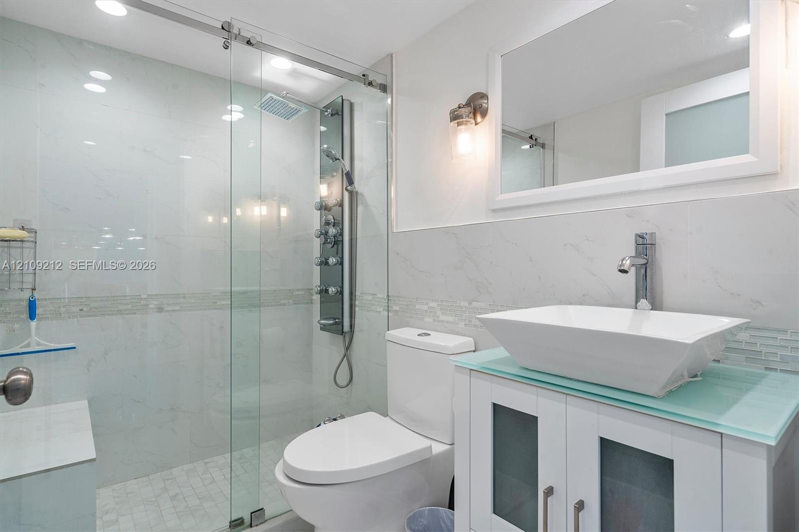 Bathroom featuring a sliding glass shower door, a multi-jet shower panel, a vessel sink with a glass countertop, and a vanity with frosted glass cabinet inserts