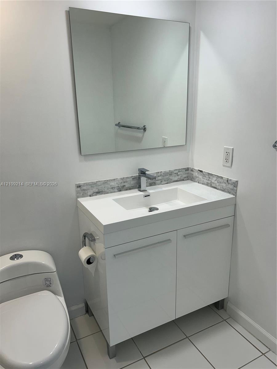 Modern vanity featuring a white finish, stone-composite countertop, integrated sink, and polished chrome hardware, complemented by a stone-tile backsplash and a frameless mirror