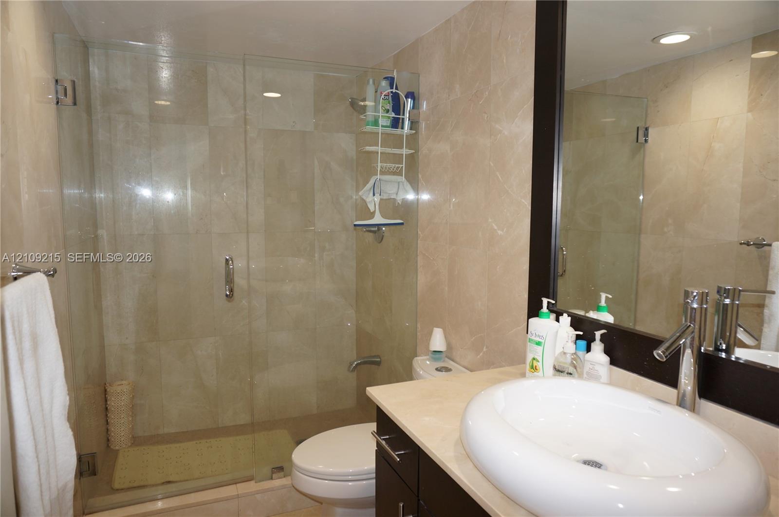 Full-wall stone tiling, glass-enclosed shower, dark-finish cabinetry, countertop-mounted oval sink, and framed mirror