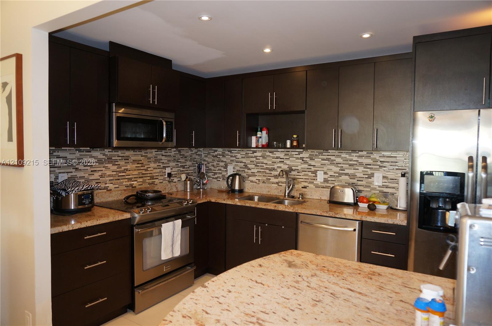 Kitchen featuring dark-toned cabinetry, light-toned stone countertops, multi-tonal mosaic tile backsplash, recessed lighting, and stainless steel appliances