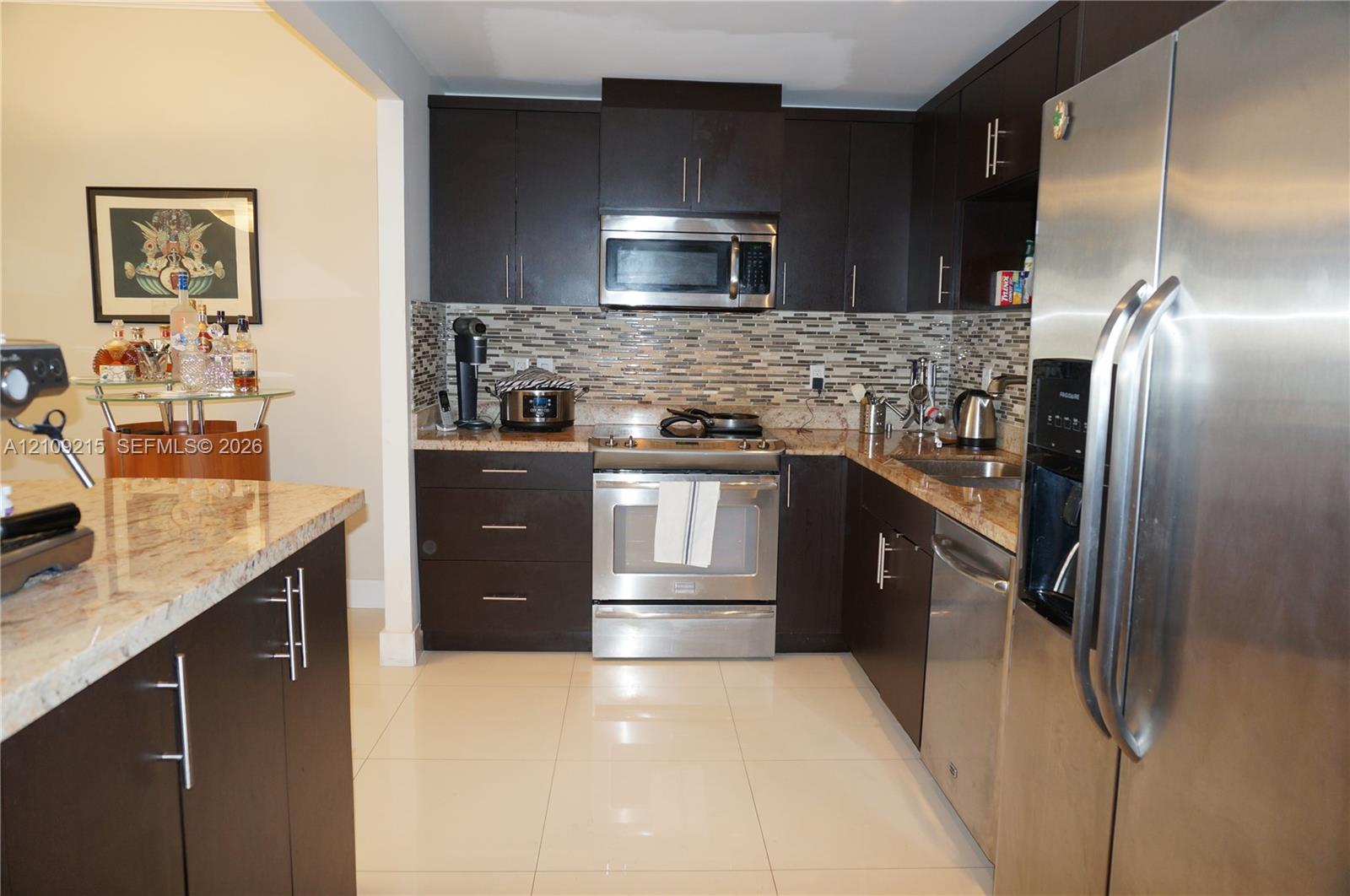Kitchen featuring dark wood-finish cabinetry, stone countertops, stainless steel appliances, multi-toned tile backsplash, and light-colored flooring