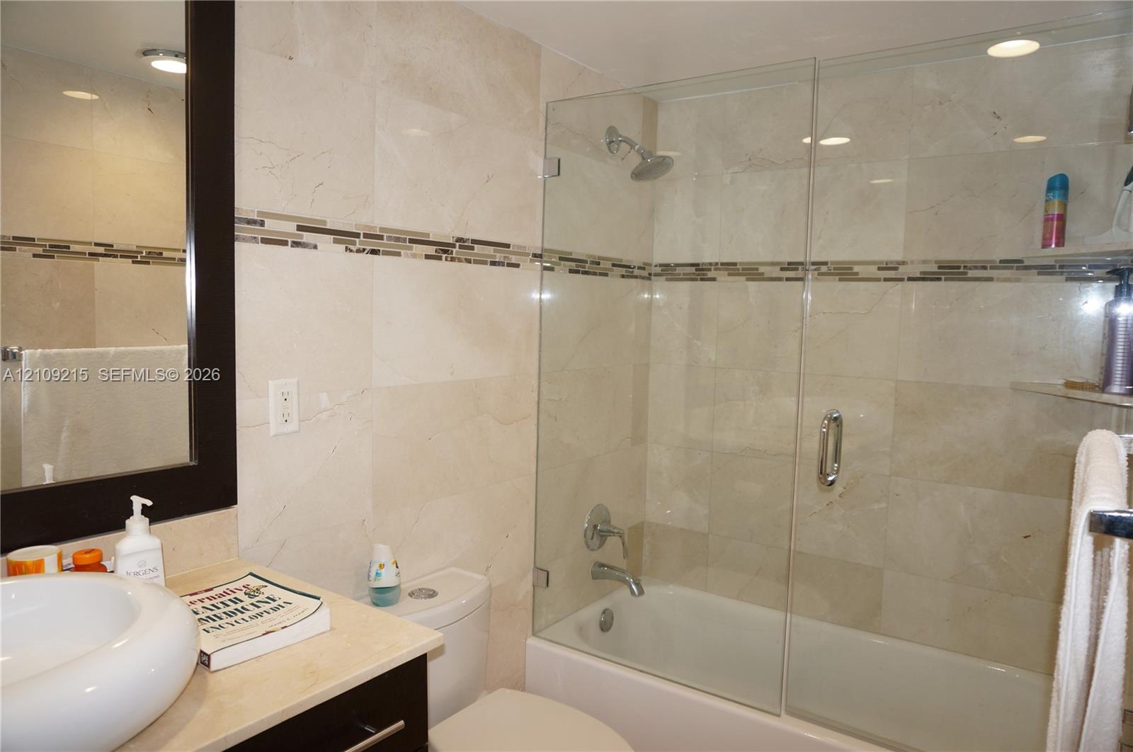 Bathroom featuring full-height stone wall tiling with decorative glass mosaic inlay, a glass-enclosed shower and tub combination, a circular vessel sink, and an integrated dark-finish vanity