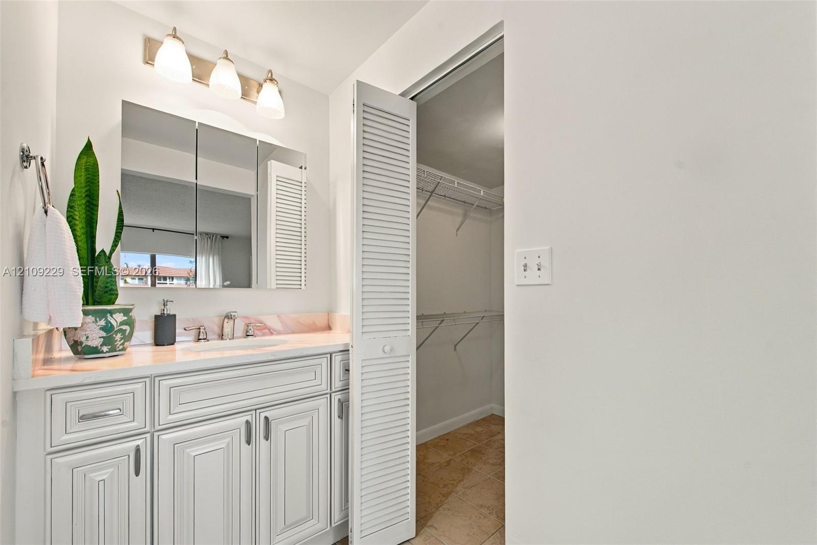 Vanity featuring white recessed-panel cabinetry, integrated stone-finish countertop with backsplash, three-light wall fixture, and tri-fold mirrored medicine cabinet, paired with a louvered-door walk-in closet