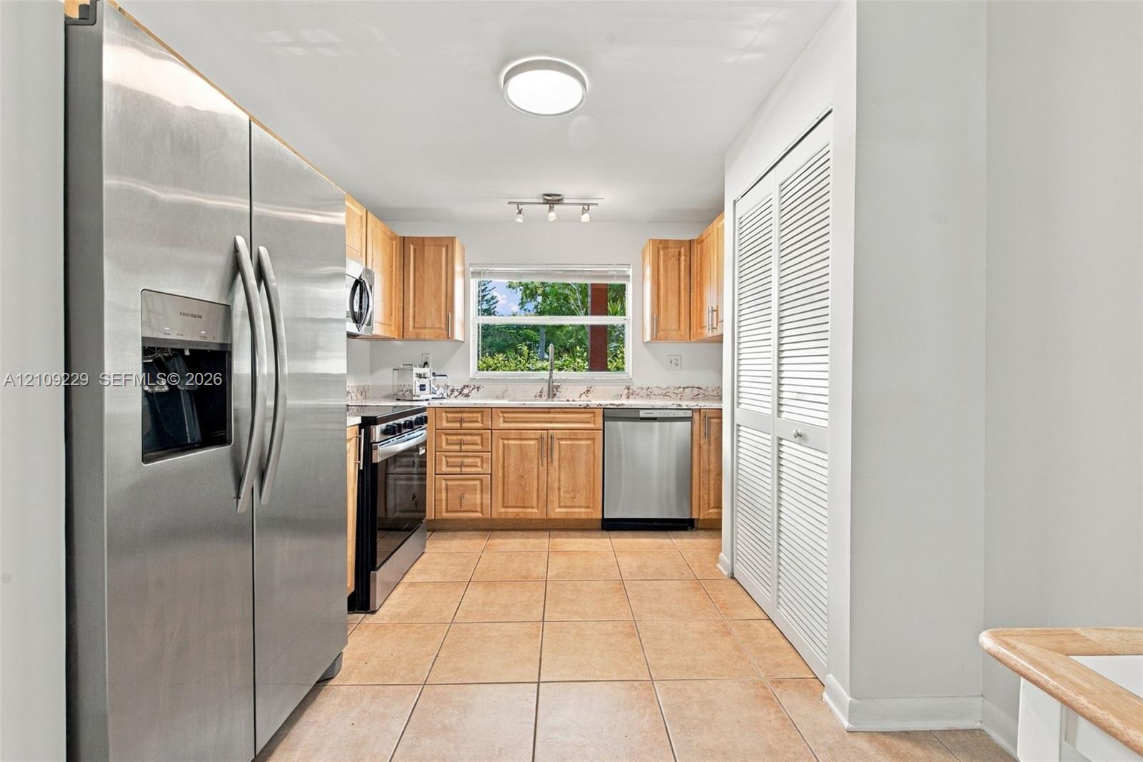 Kitchen featuring light-finish wood cabinetry, stone countertops, stainless steel appliances, ceramic tile flooring, and louvered double-door storage Kitchen featuring light-finish wood cabinetry, stone countertops, stainless steel appliances, ceramic tile flooring, and louvered double-door storage
