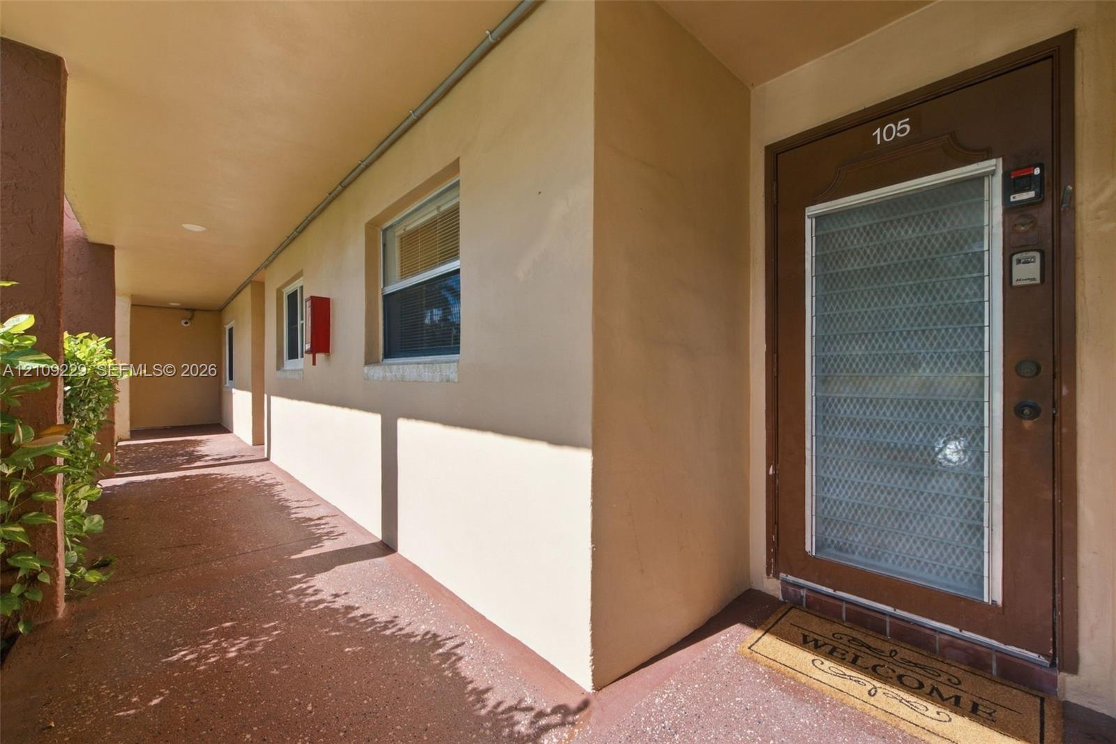 Covered exterior walkway with textured flooring, smooth stucco exterior, and a metal security door featuring an integrated glass panel Covered exterior walkway with textured flooring, smooth stucco exterior, and a metal security door featuring an integrated glass panel
