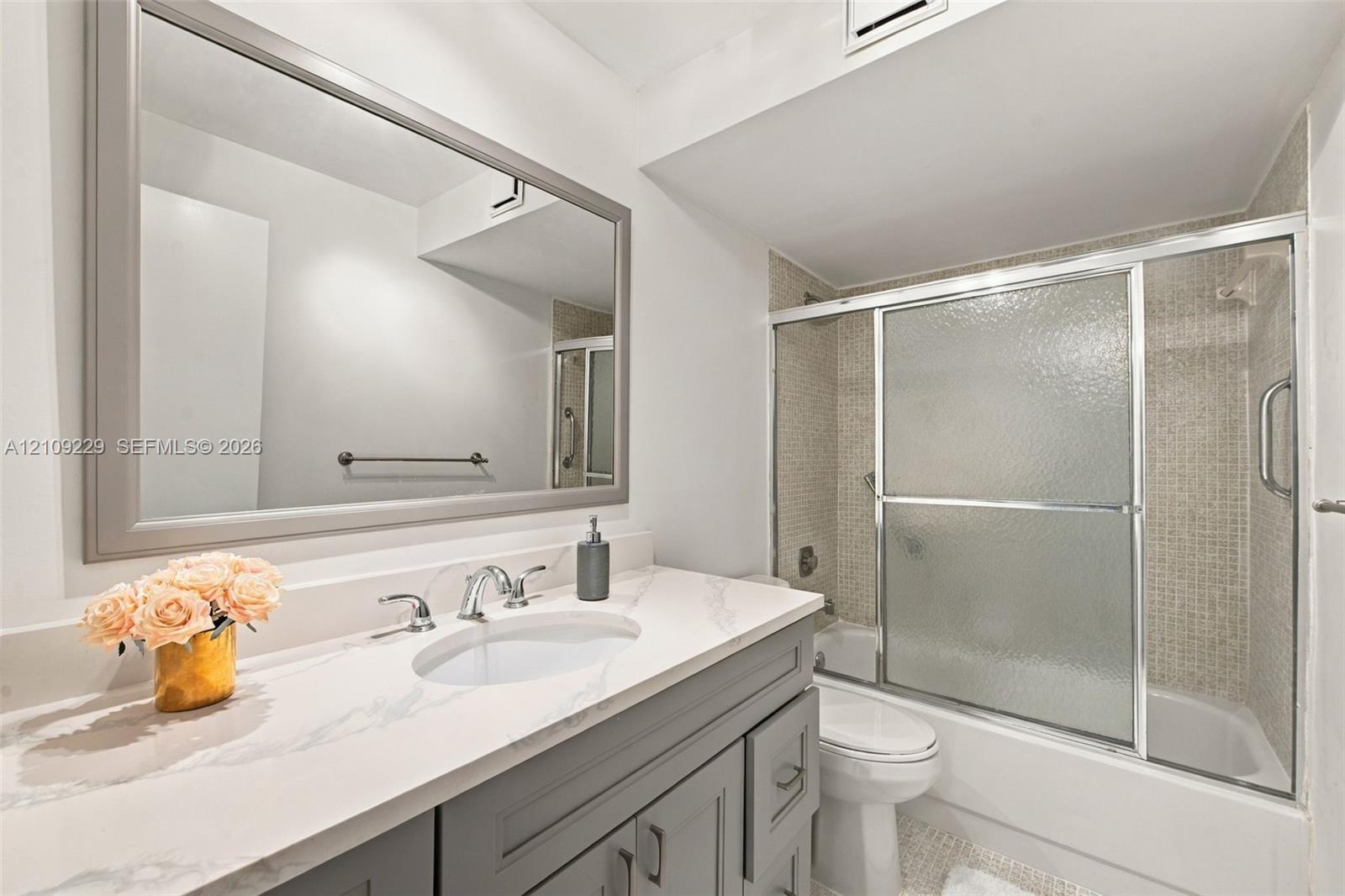 Bathroom featuring a vanity with a light-toned stone countertop, undermount sink, and brushed metal faucet, paired with a large framed mirror and a bathtub-shower combination with sliding glass doors