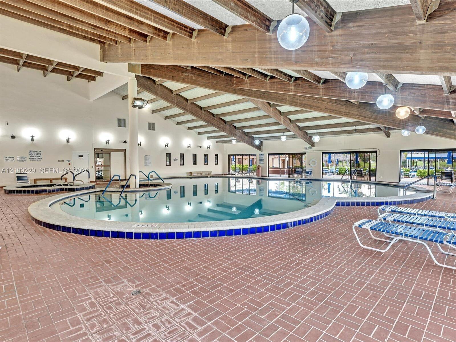 Indoor swimming pool featuring an expansive free-form basin, an integrated spa, exposed wood ceiling beams, pendant globe lighting, and brick flooring