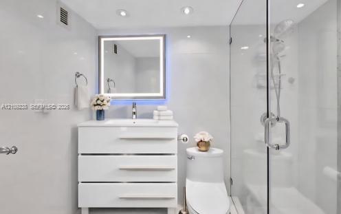 Bathroom featuring a white three-drawer vanity, illuminated rectangular mirror, one-piece elongated toilet, and a glass-enclosed shower with polished chrome fixtures