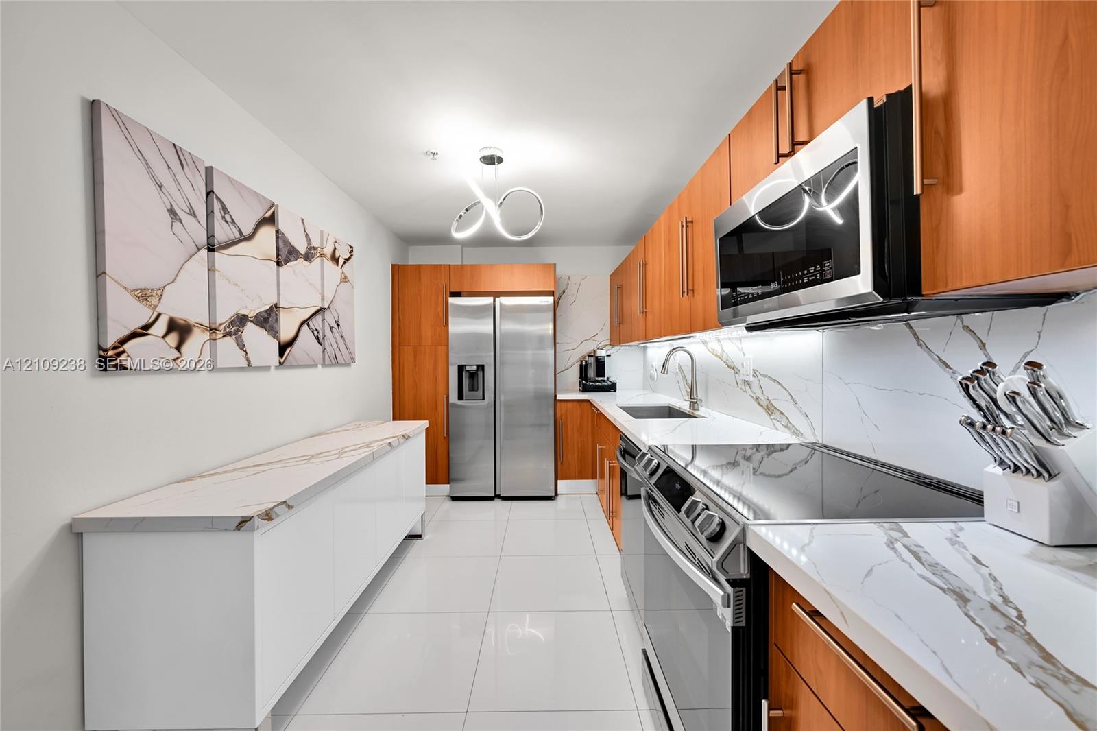 Galley kitchen featuring wood cabinetry, stone-surface countertops with matching backsplashes, stainless steel appliances, polished tile flooring, and a contemporary architectural light fixture
