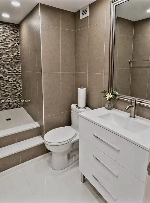 Bathroom featuring a modern white vanity, glass-tiled shower wall accent, large-format neutral wall tiling, stepped shower entry, and a framed mirror