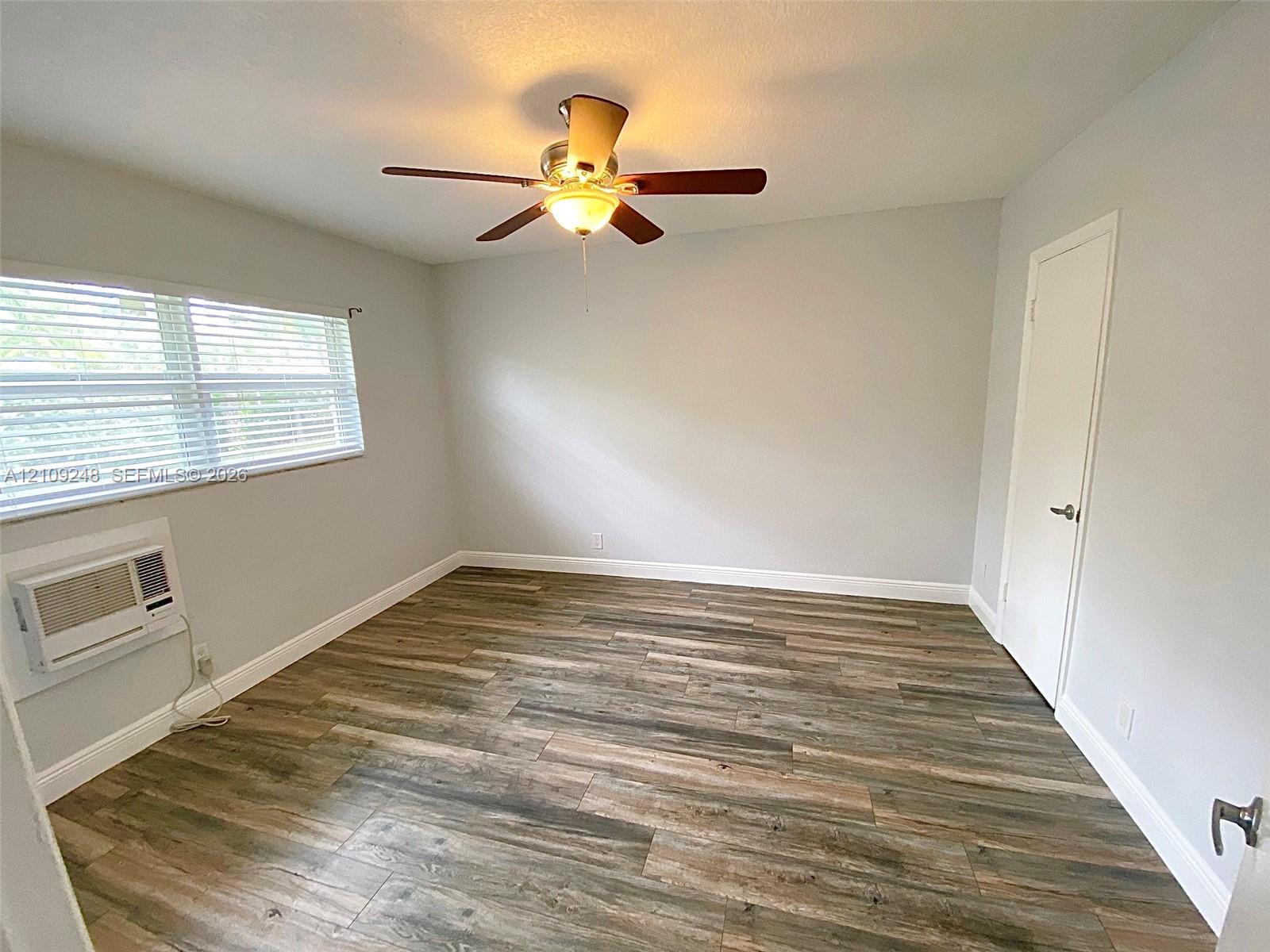 Room featuring wood-finish flooring, a ceiling fan with integrated lighting, horizontal window blinds, and a wall-mounted cooling unit