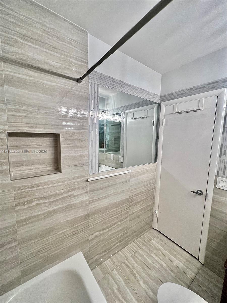 Bathroom featuring neutral-toned stone-finish wall and floor tile, recessed wall niche, glass-fronted medicine cabinet, and decorative linear mosaic tile trim
