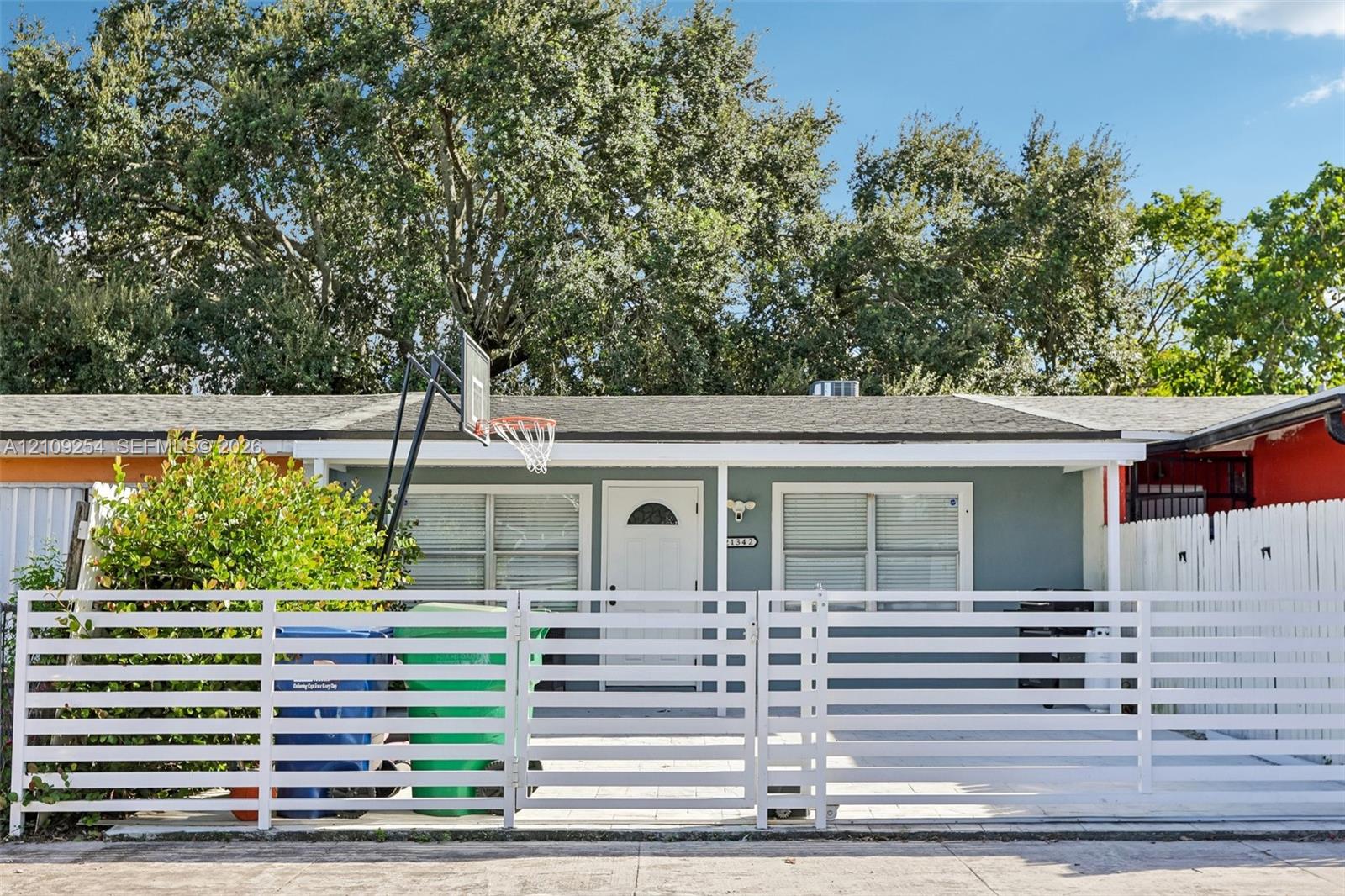 Single-story residence featuring a gray exterior, a covered front entryway, horizontal white slat fencing, and mature trees Single-story residence featuring a gray exterior, a covered front entryway, horizontal white slat fencing, and mature trees
