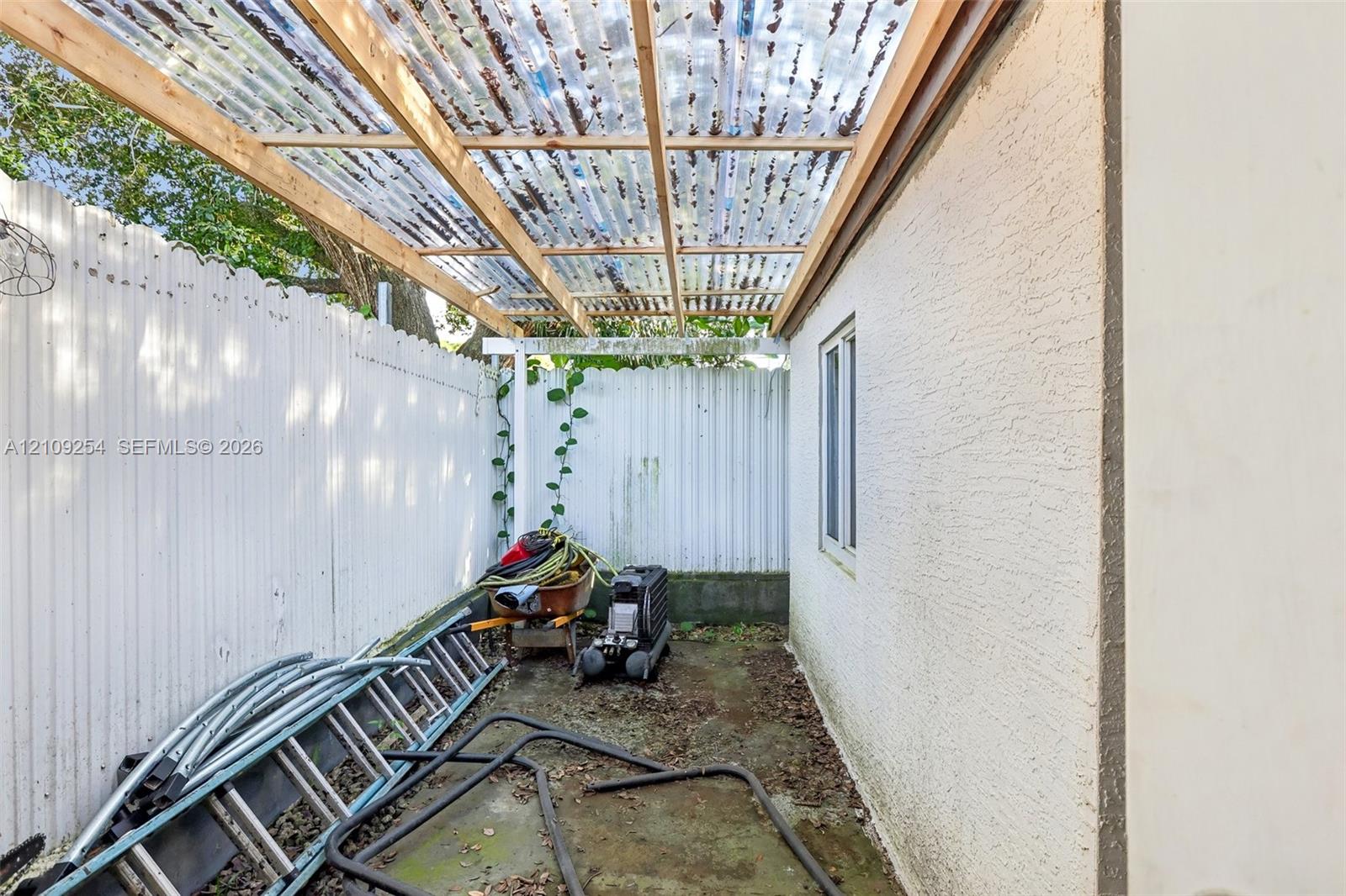 Side patio featuring a corrugated translucent roof, wood framing, stucco exterior wall, and white privacy fencing