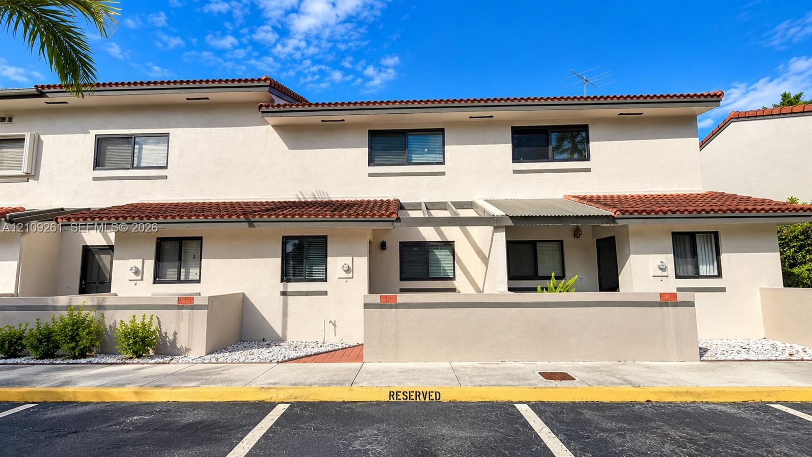 Two-story exterior featuring light-toned stucco, a terracotta tile roof, structural wooden pergola, and low-profile walled front courtyards