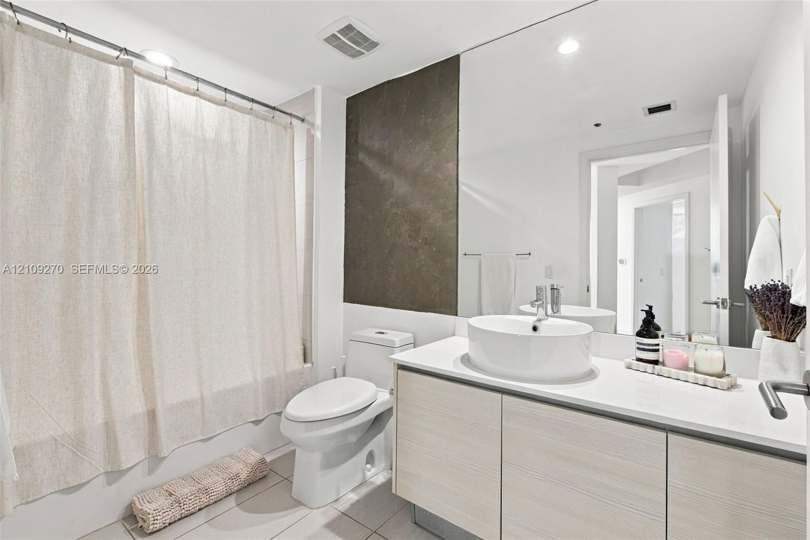 Bathroom featuring a vanity with wood-finish cabinetry, a white countertop, and a vessel sink, accompanied by a full-wall mirror, tiled flooring, and a tub-shower combination with a large-format tiled wall accent