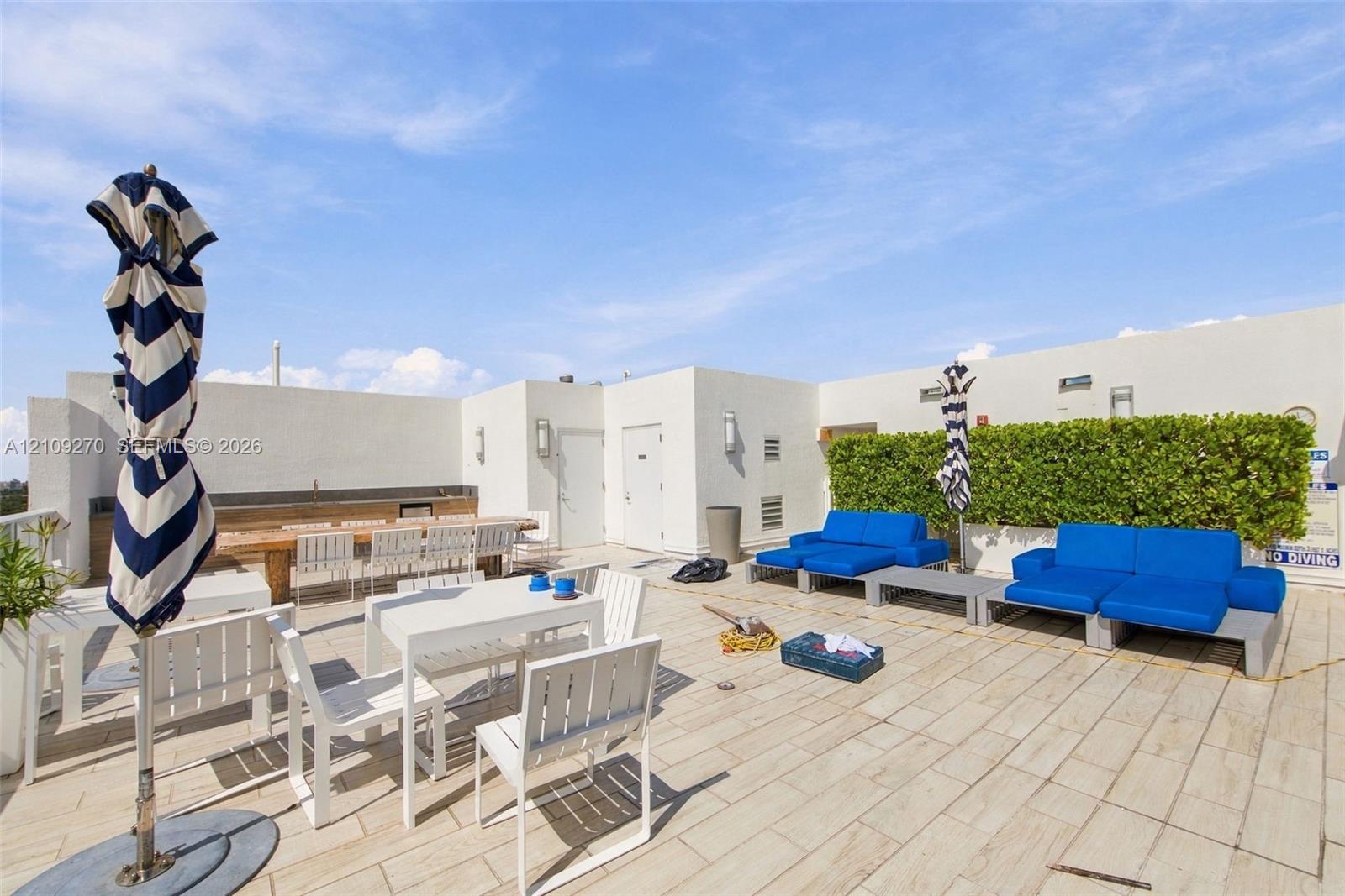 Rooftop terrace featuring light-toned tile flooring, integrated perimeter hedge, stucco wall finishes, and built-in outdoor kitchen structure with attached wall-mounted lighting
