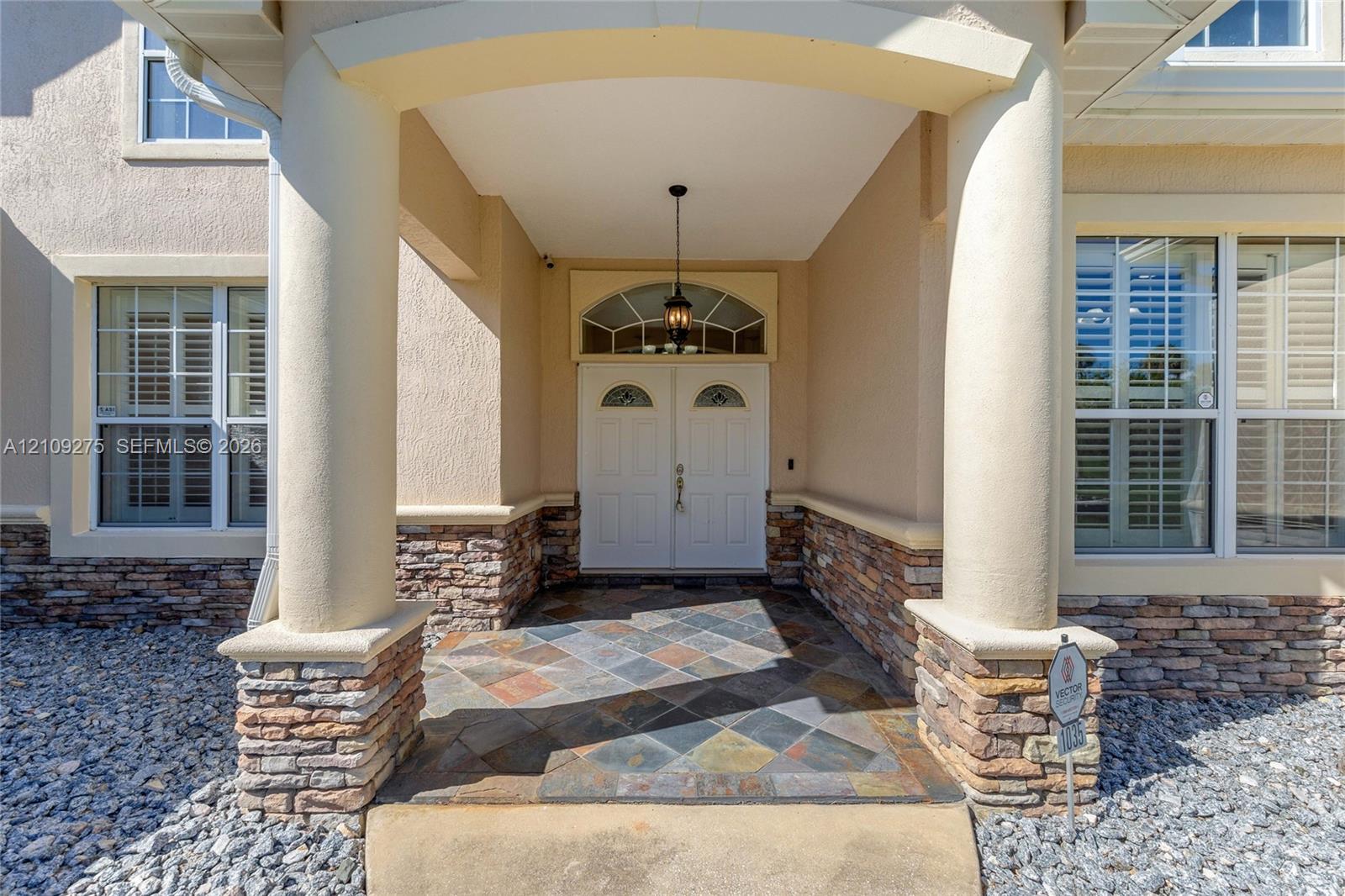 Covered entryway featuring prominent structural columns, stone veneer accents, multi-tonal slate tile flooring, and double front doors with an arched transom window