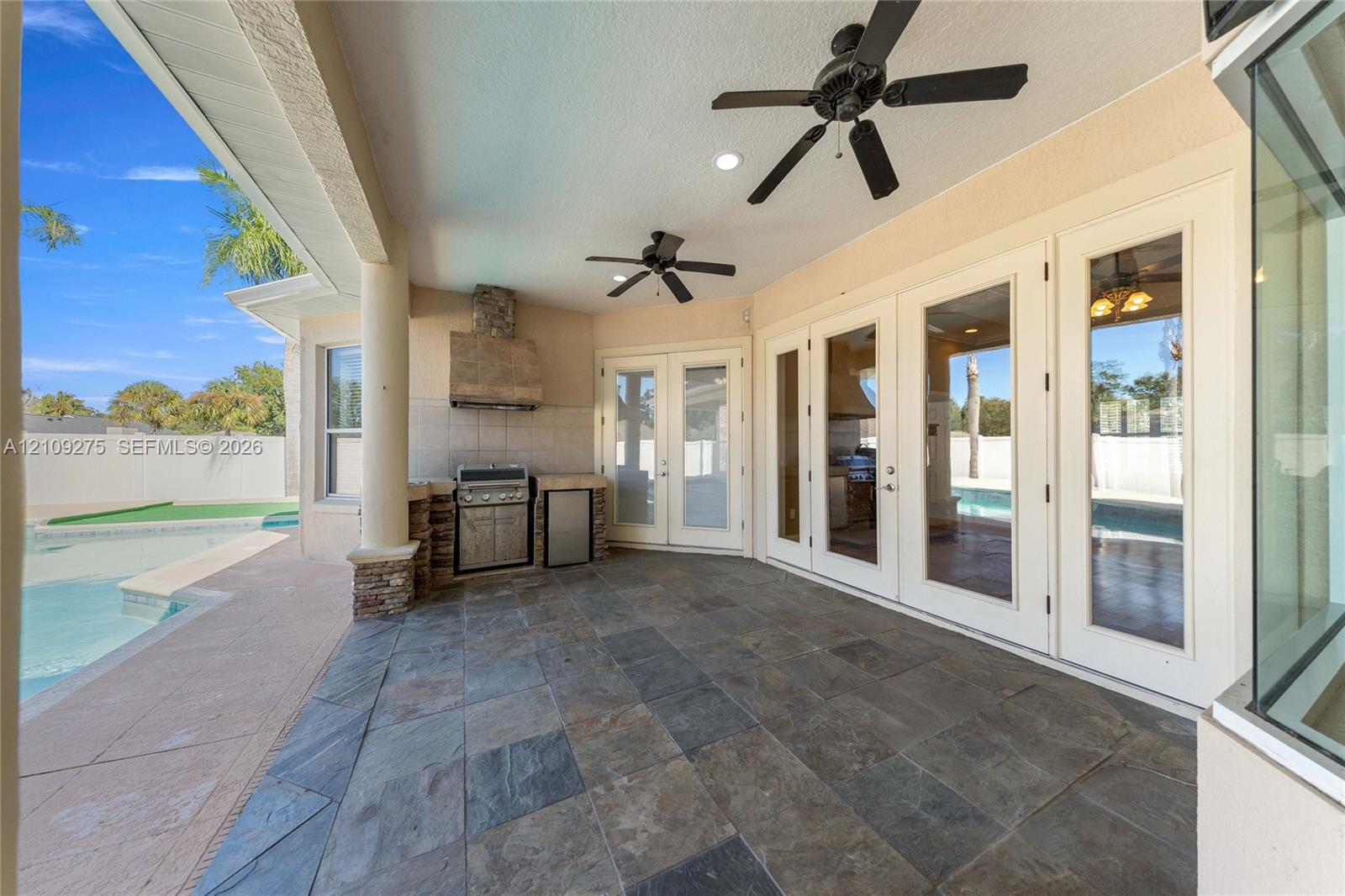 Covered patio featuring stone-tile flooring, built-in outdoor kitchen with stainless steel grill, recessed ceiling lighting, dual overhead fans, and multi-panel glass entry doors