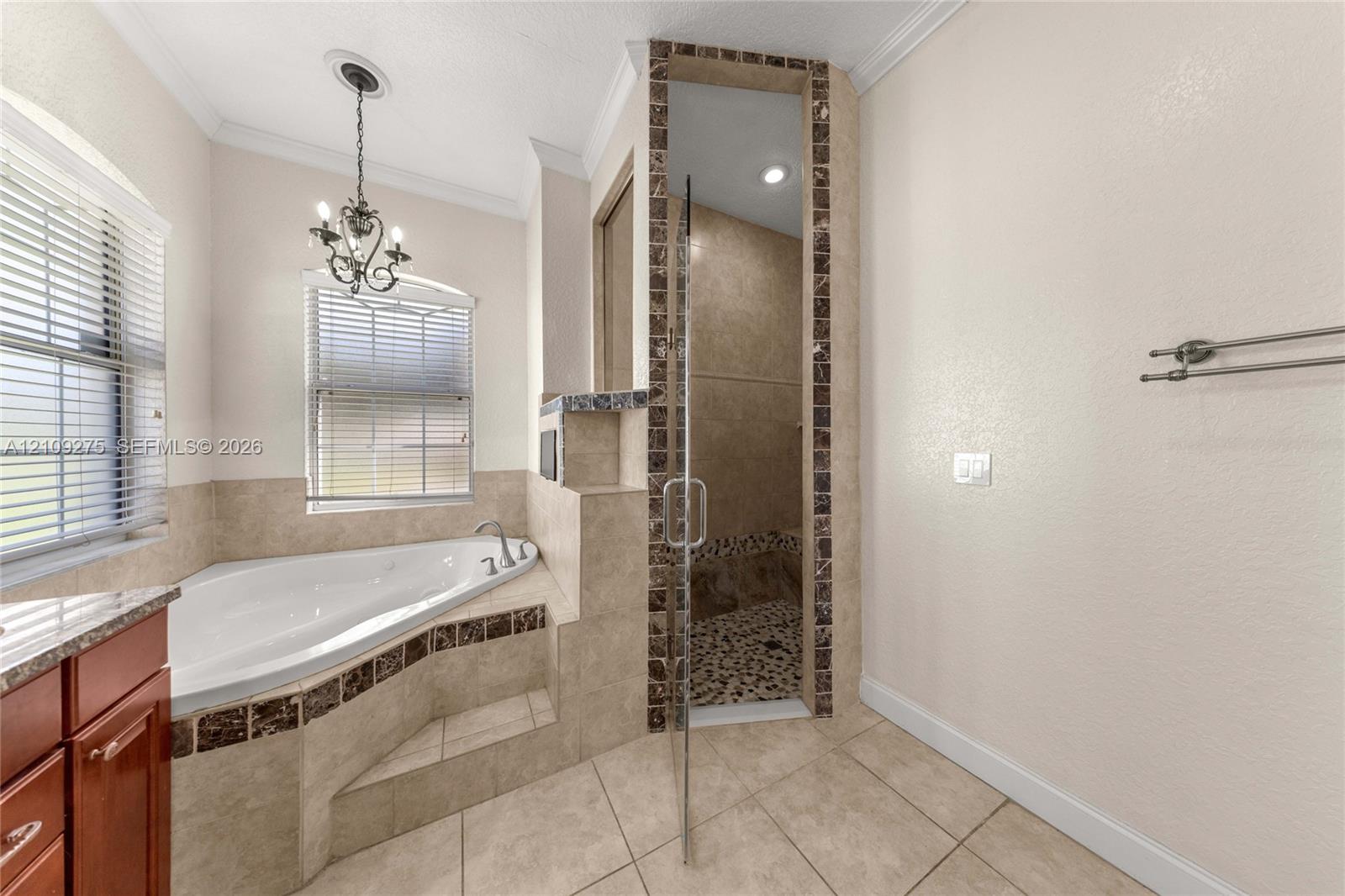 Corner jetted soaking tub featuring decorative tile accents, glass-enclosed shower with stone tile flooring, crown molding, and metal chandelier fixture