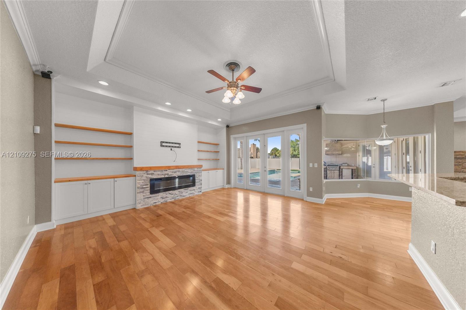 Living area featuring wood-finish flooring, tray ceiling, built-in shelving, stone-finish fireplace, and multi-panel glass door