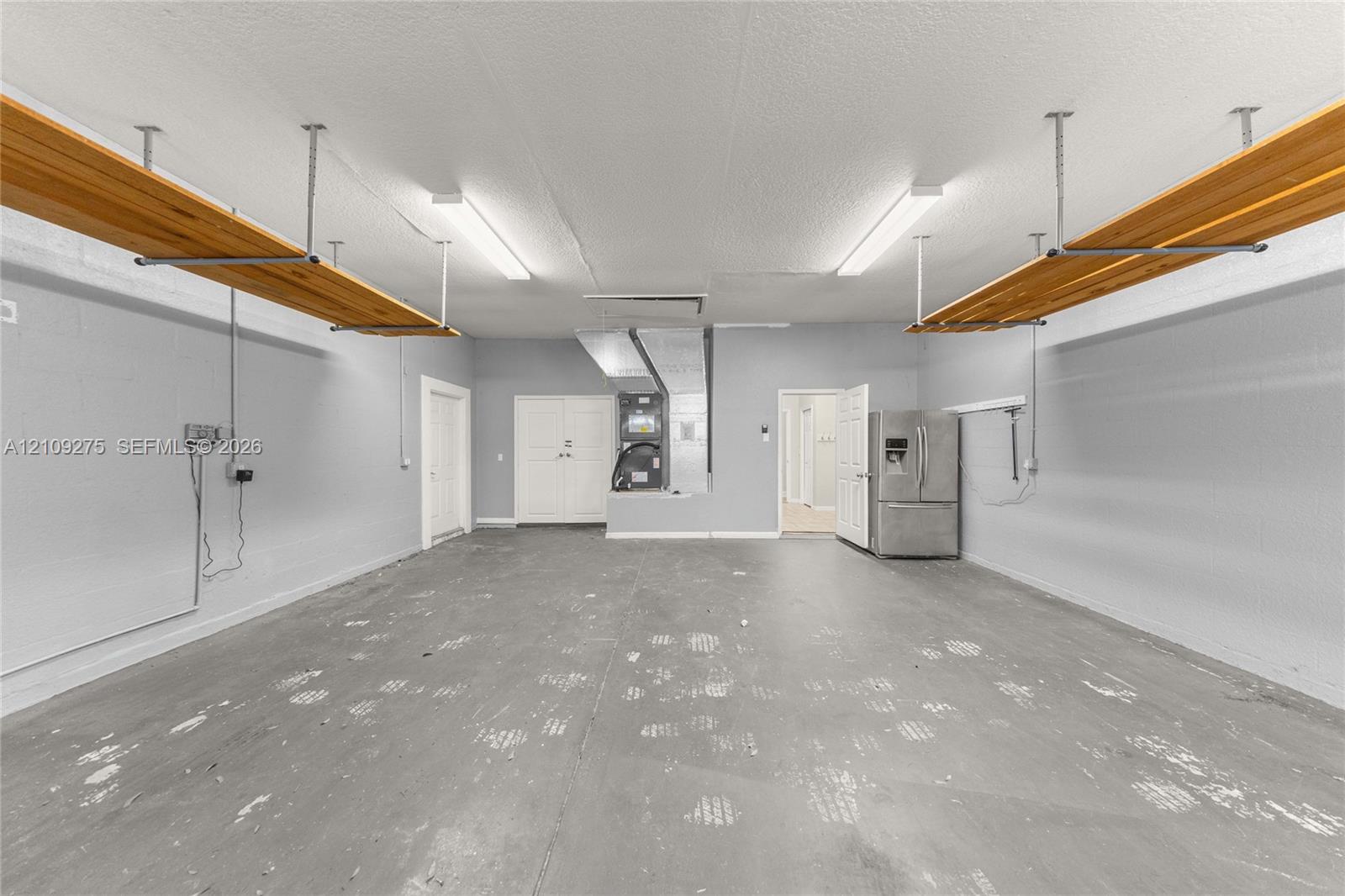 Spacious garage featuring high-capacity overhead wooden shelving, dual linear ceiling-mounted lighting fixtures, finished gray walls, and painted concrete flooring