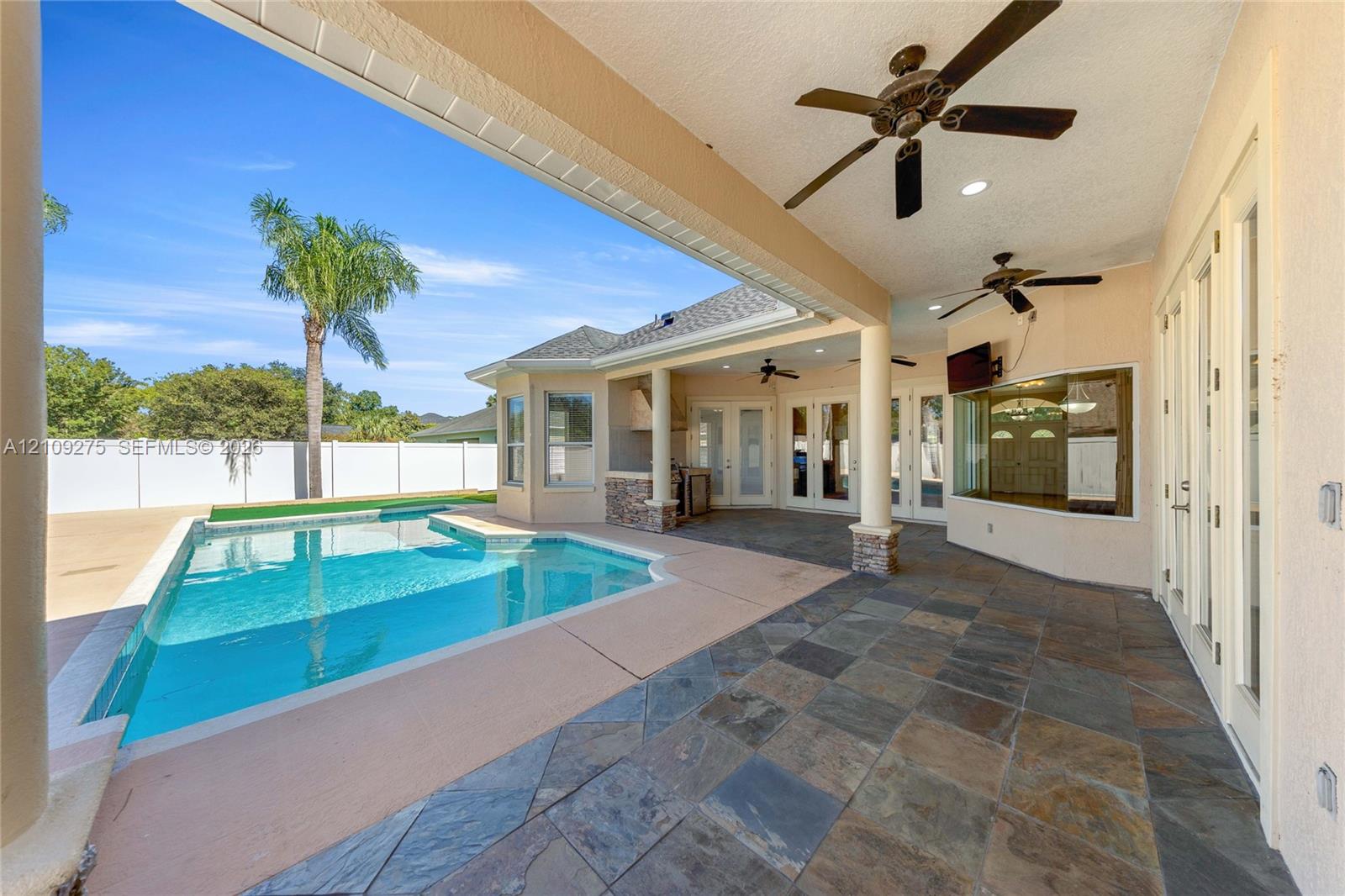 Covered patio featuring stone-tile flooring, multiple ceiling fans, recessed lighting, and a built-in outdoor kitchen, opening to a swimming pool with a concrete deck and privacy fencing