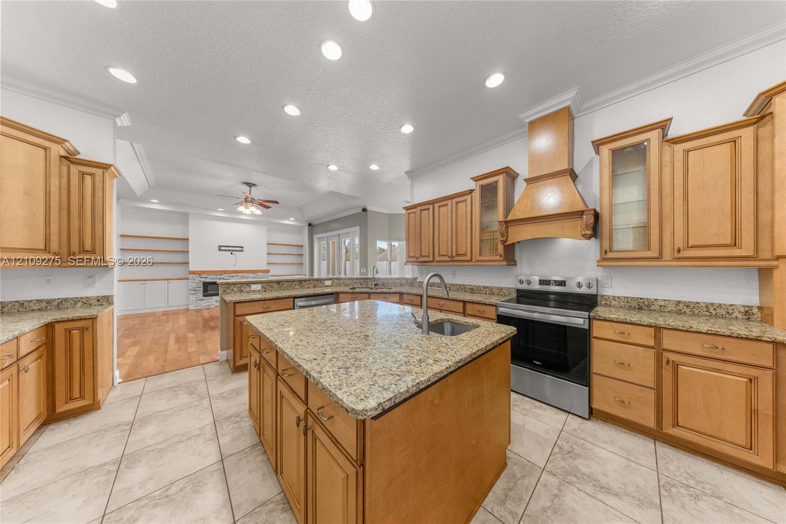 Kitchen featuring wood cabinetry, stone countertops, a center island with an integrated sink, a custom wood range hood, and recessed lighting