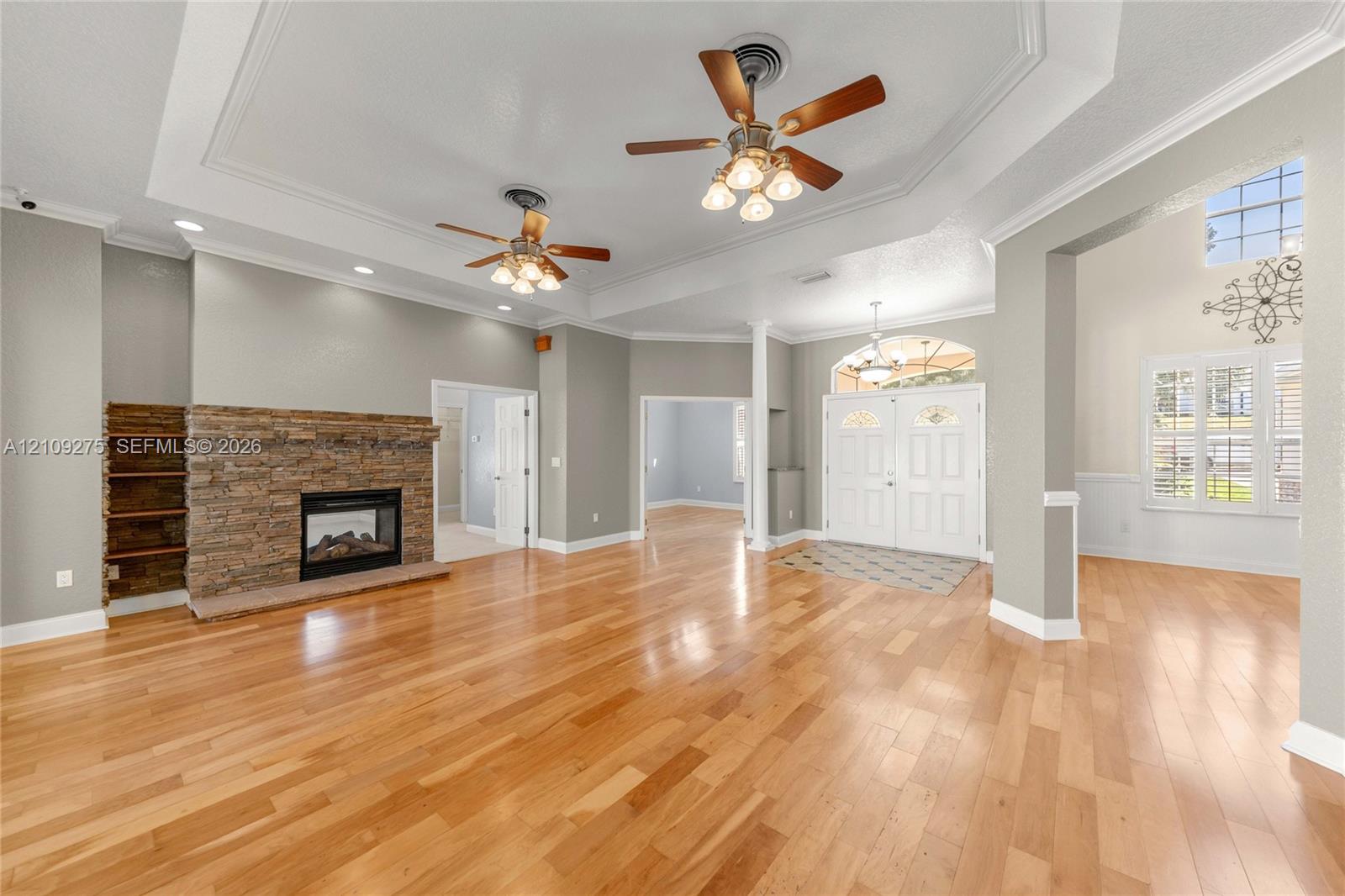Spacious interior featuring wood-finish flooring, a stone-faced fireplace with built-in shelving, tray ceilings, and dual ceiling fans