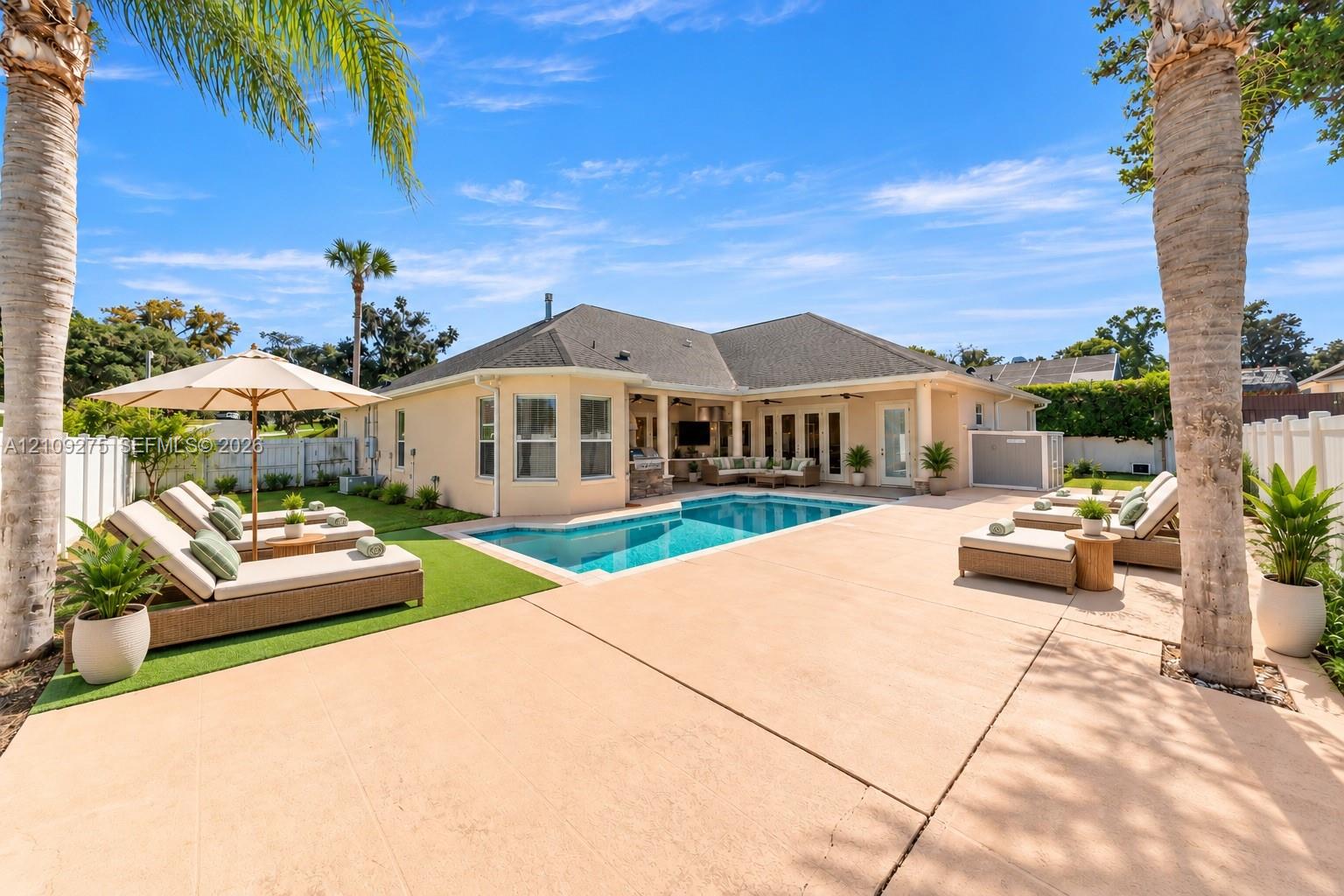 Backyard oasis featuring an in-ground pool, expansive concrete patio, covered lanai, synthetic turf lawn, and mature palm trees