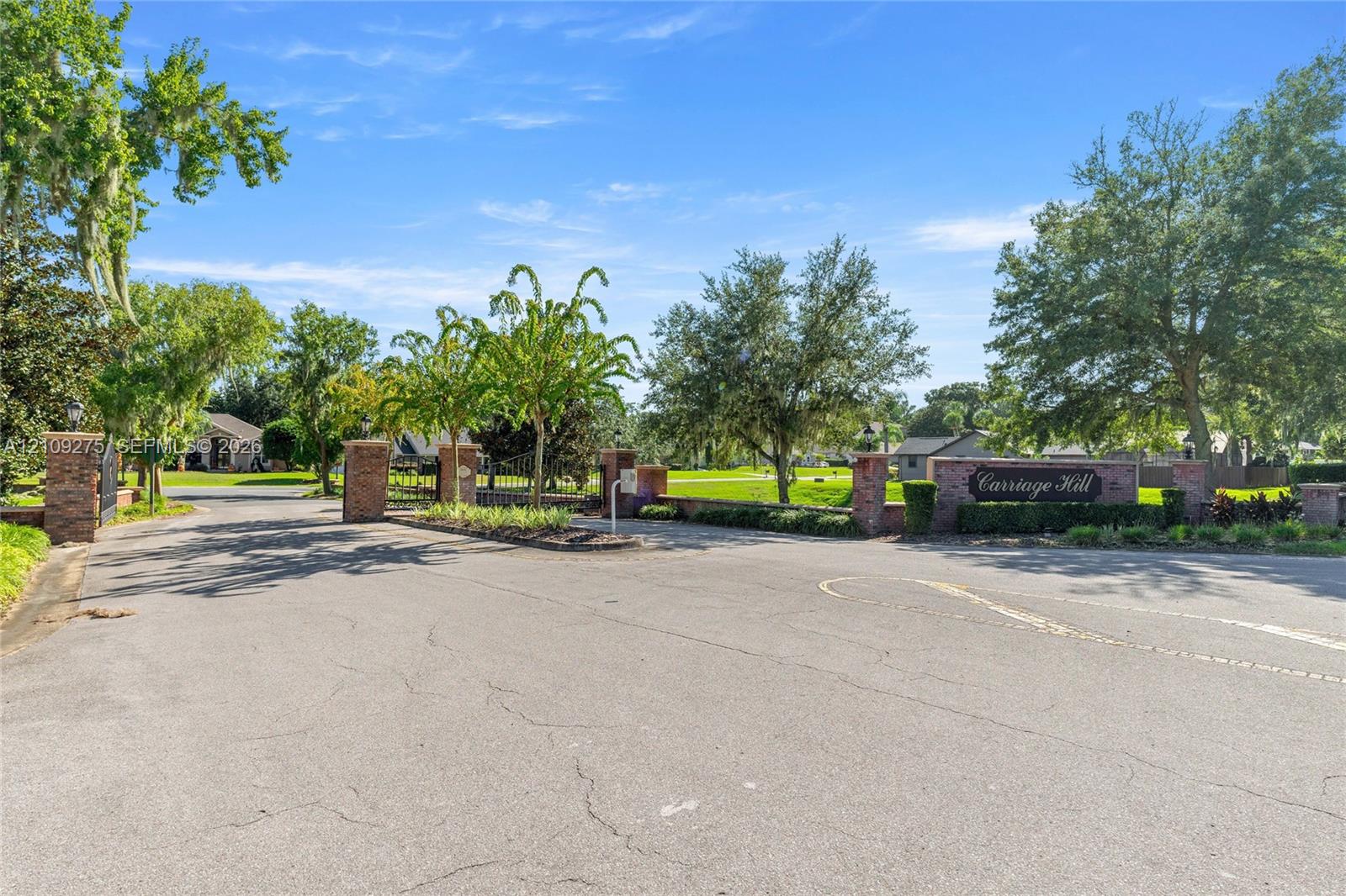 Entryway featuring brick masonry pillars, ornamental metal gate, paved access road, and mature landscaping