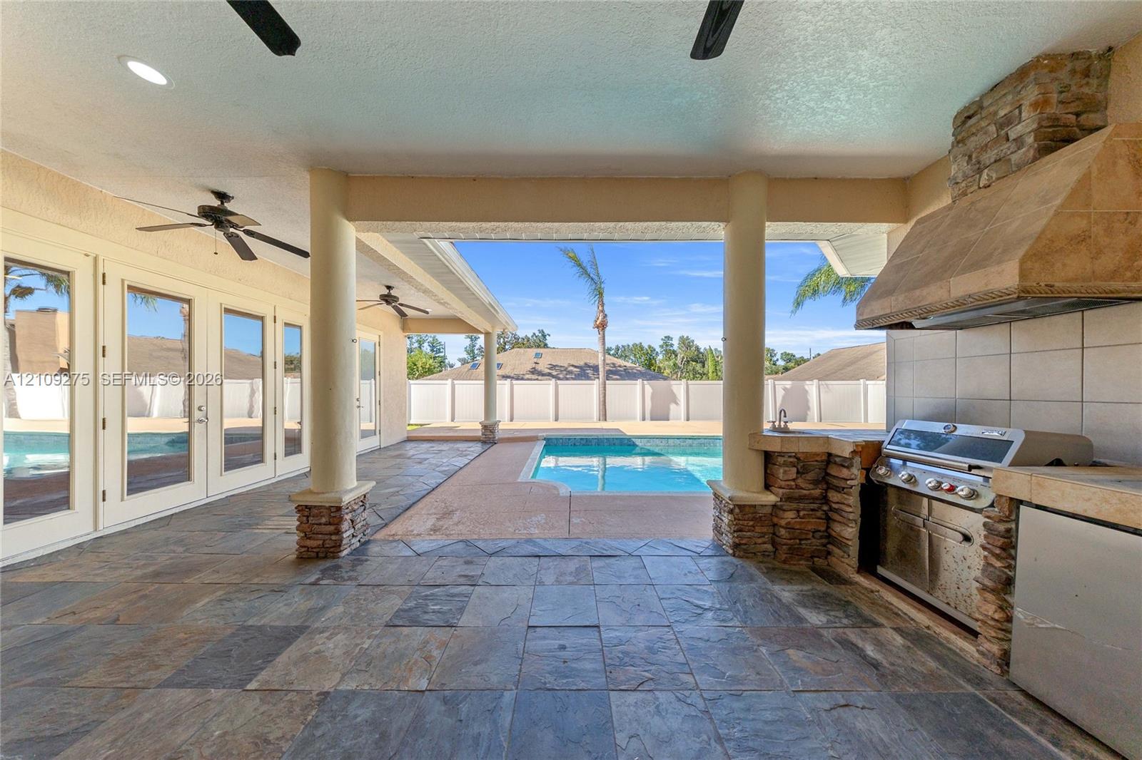 Covered patio featuring slate-style tile flooring, built-in outdoor kitchen with stone veneer base and tiled hood, columns with stone accents, and an integrated swimming pool