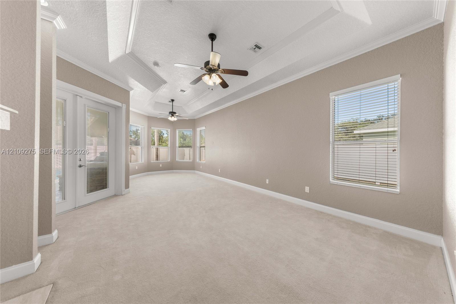 Spacious room featuring a tray ceiling, dual ceiling fans with integrated lighting, neutral wall-to-wall carpeting, crown molding, and glass panel doors