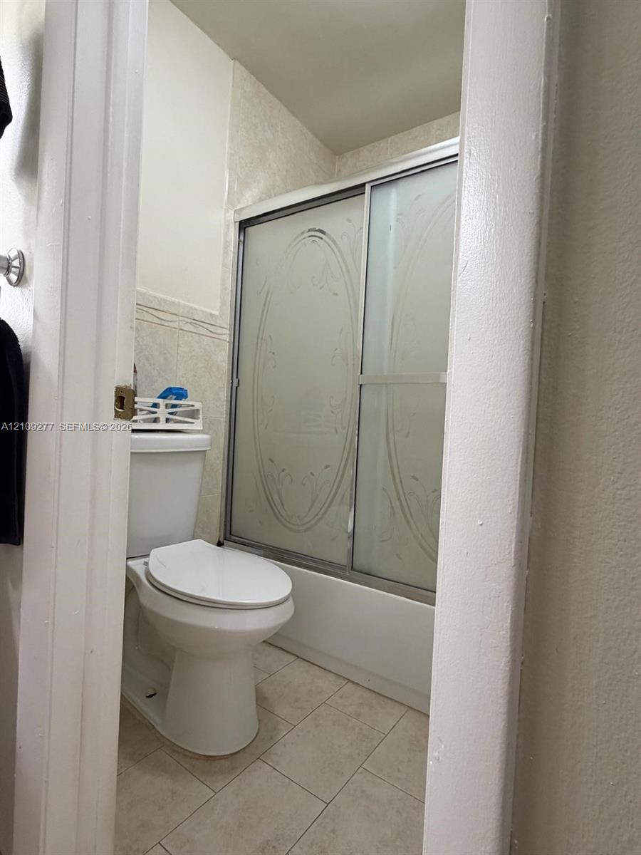Full bathroom featuring a shower-tub combination with sliding etched glass doors, tile surround, porcelain toilet, and neutral tile flooring