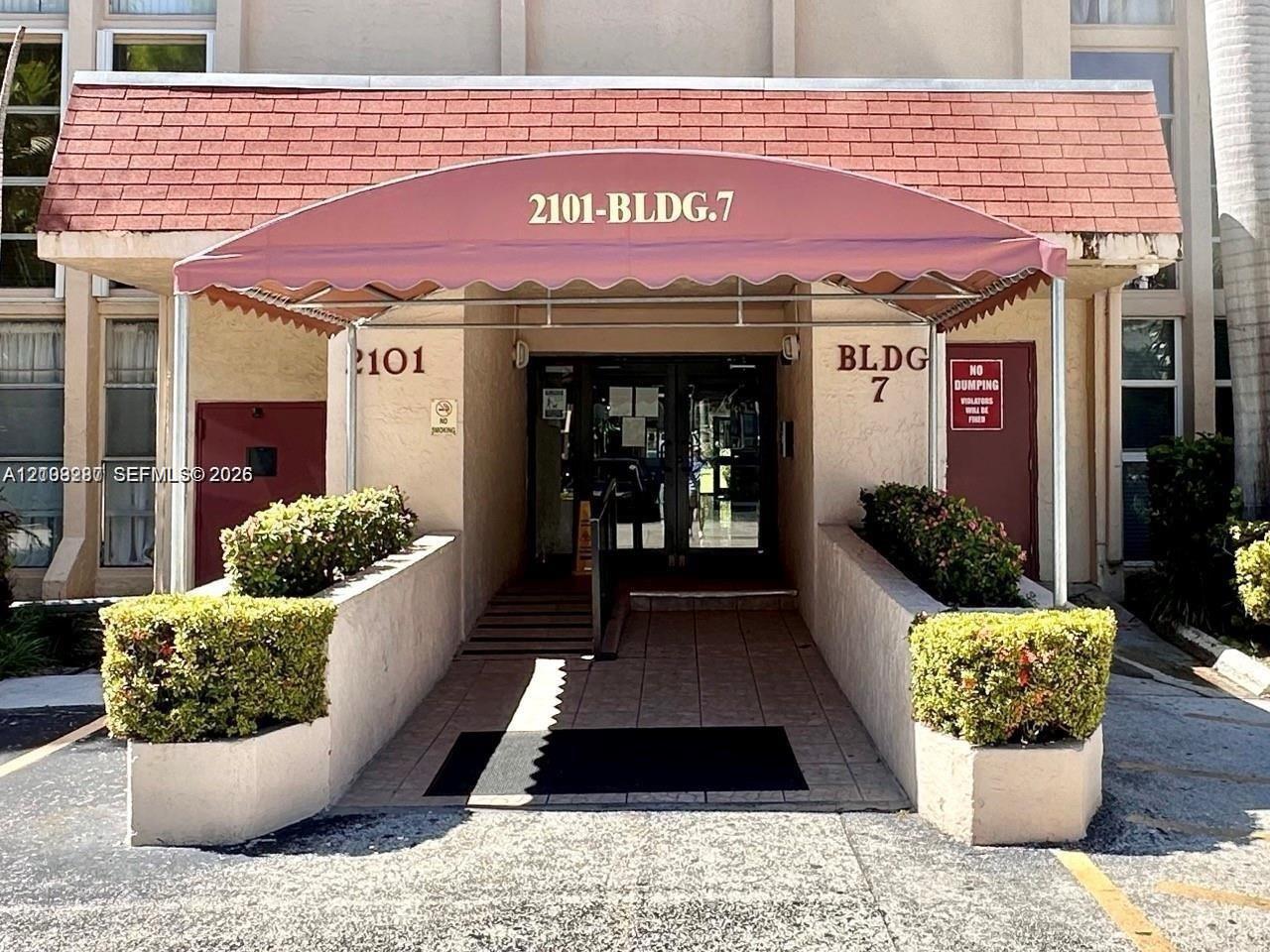 Building entrance featuring a red canvas awning, shingle-roof overhang, paved walkway, and built-in masonry planters Building entrance featuring a red canvas awning, shingle-roof overhang, paved walkway, and built-in masonry planters