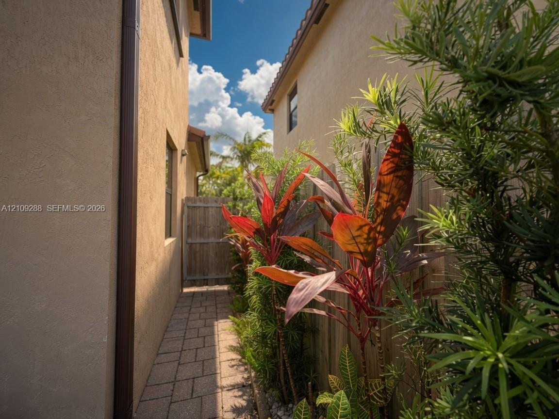 Side yard walkway featuring stone pavers, stucco exterior wall, wood-panel fencing, and mature tropical landscaping
