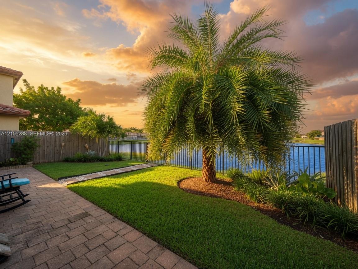 Backyard featuring a paved patio, manicured lawn, palm tree landscaping, metal fencing, and waterfront exposure