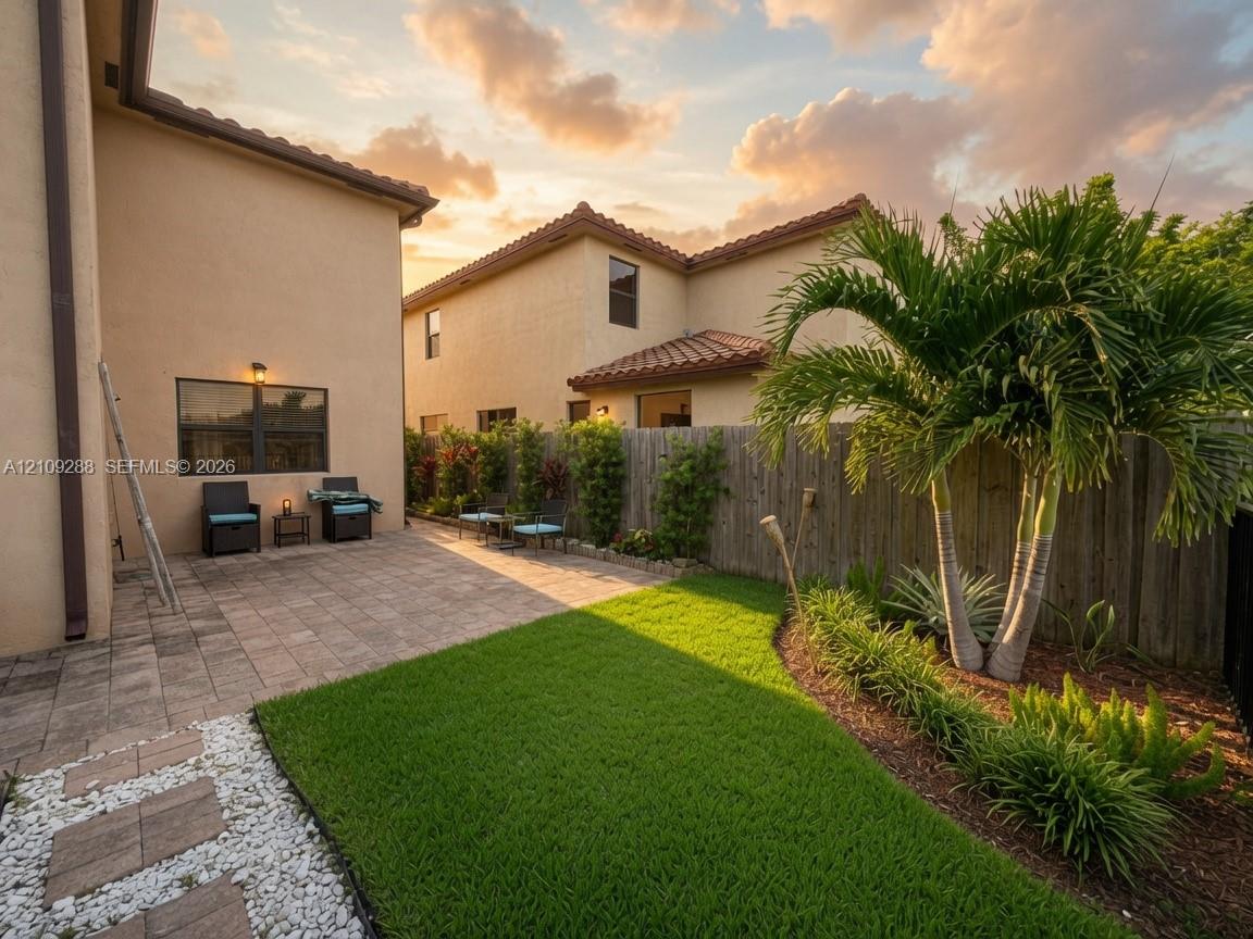 Stucco exterior featuring a paver patio, manicured lawn, wood-finish privacy fencing, and established palm landscaping