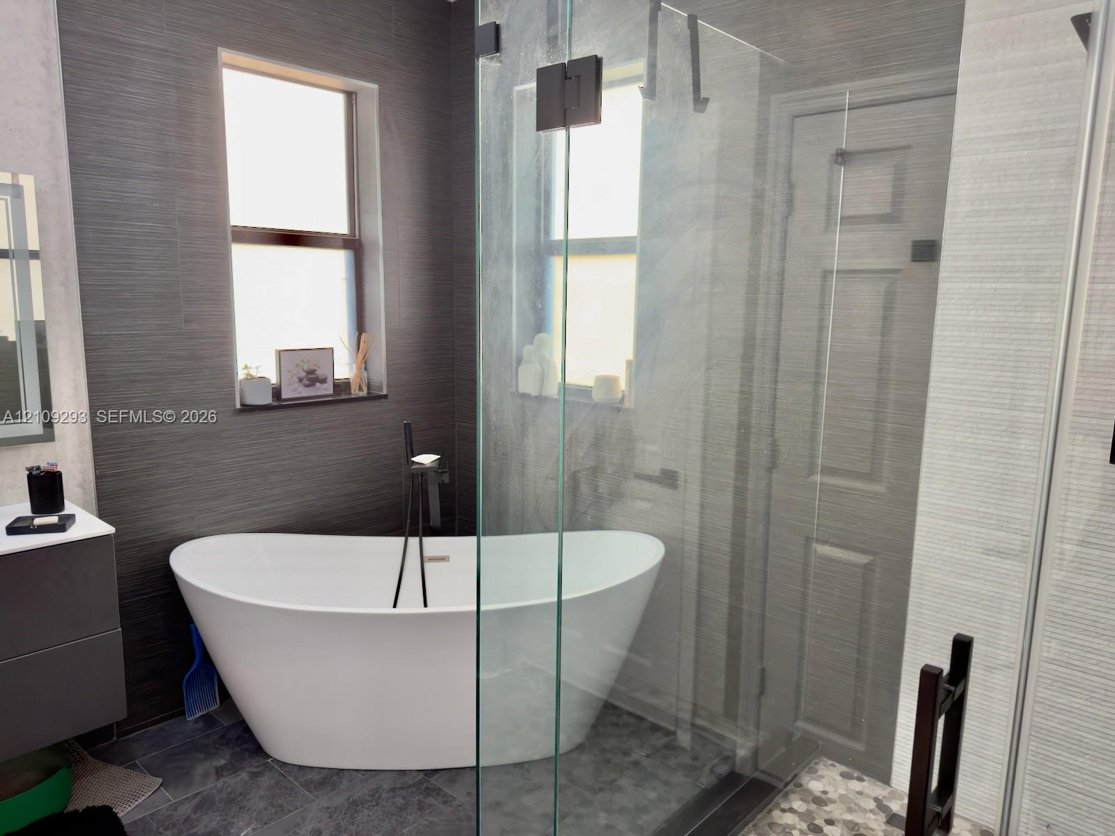 Freestanding white soaking tub with matte black floor-mounted faucet, walk-in glass-enclosed shower, dark textured wall tiling, and floating vanity
