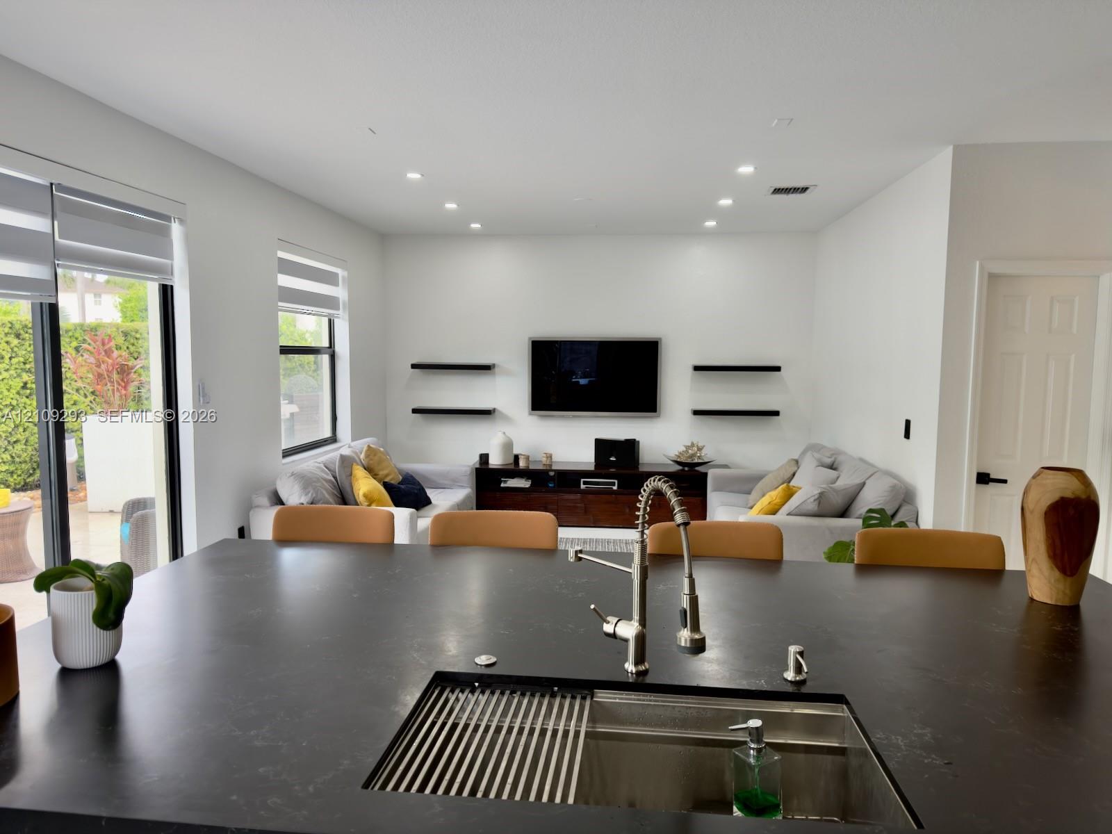 Large kitchen island featuring a dark matte countertop, undermount stainless steel sink with a high-arch pull-down faucet, recessed ceiling lighting, and wall-mounted floating shelves