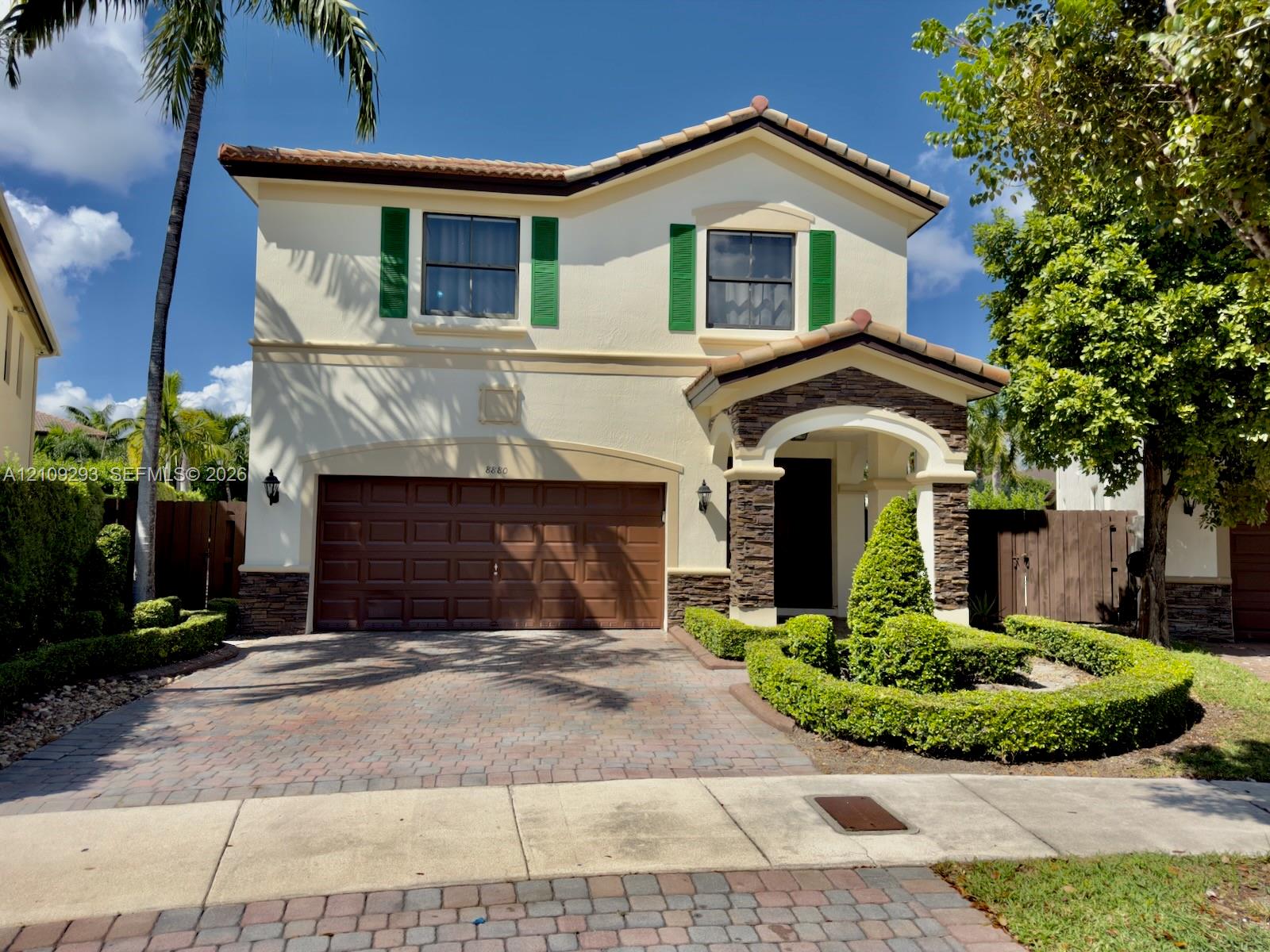 Stucco exterior featuring decorative green shutters, stone-veneer accents, a paved driveway, and manicured landscaping