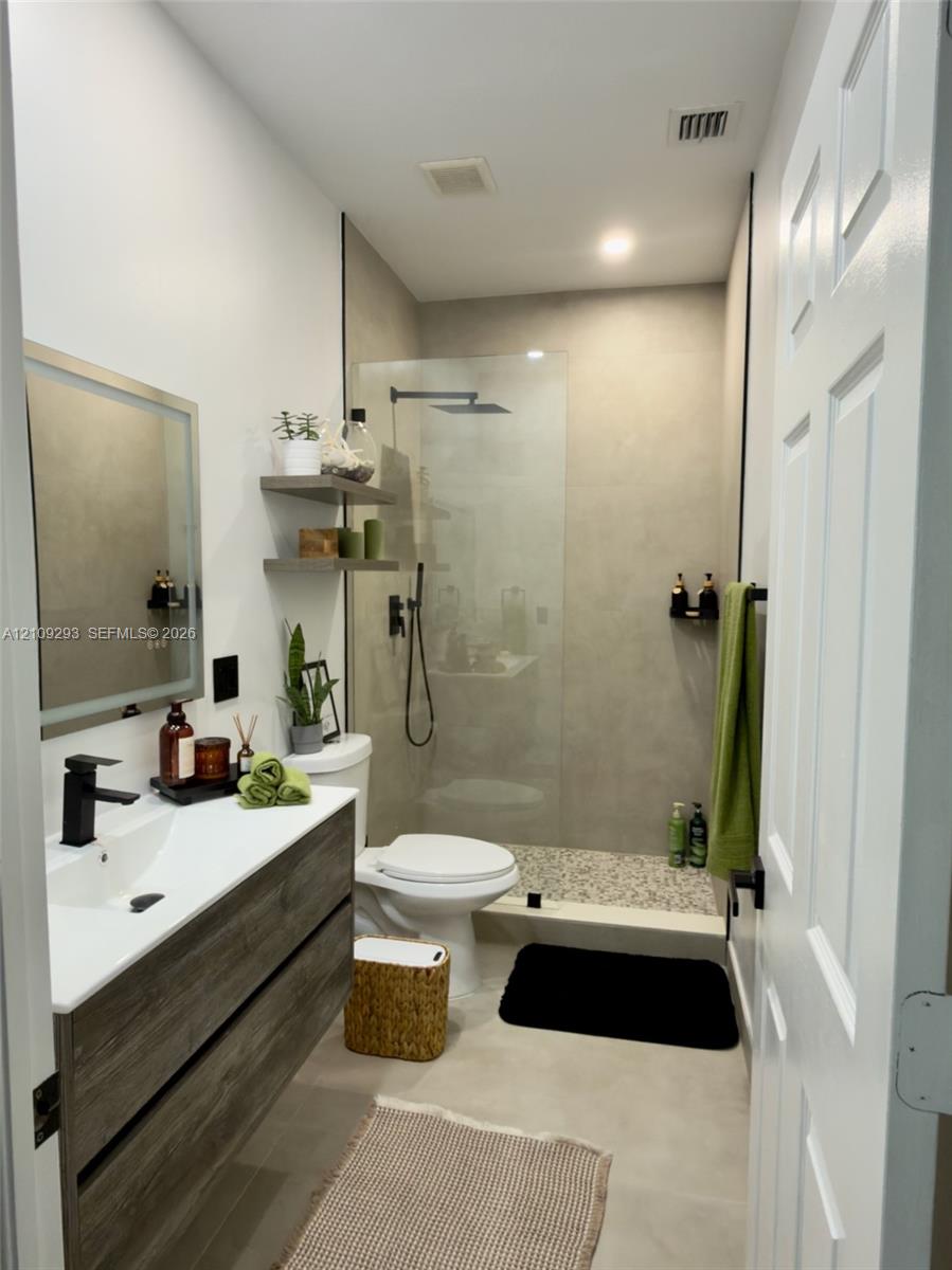 Floating wood-finish vanity with a white integrated sink, matte black faucet, illuminated mirror, walk-in shower with matte black rainfall fixture, and neutral large-format tile flooring