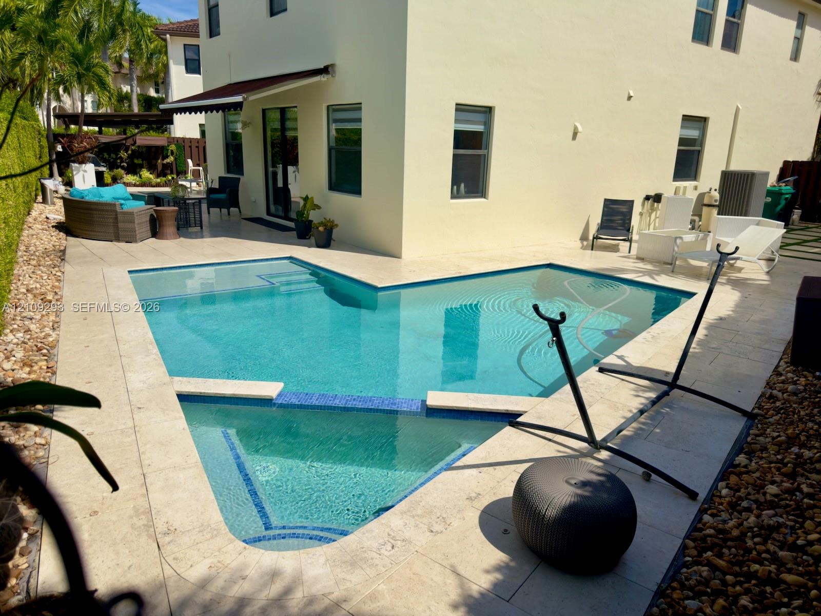 Custom-shaped swimming pool with blue tile accents, expansive stone paver pool deck, and retractable wall-mounted awning