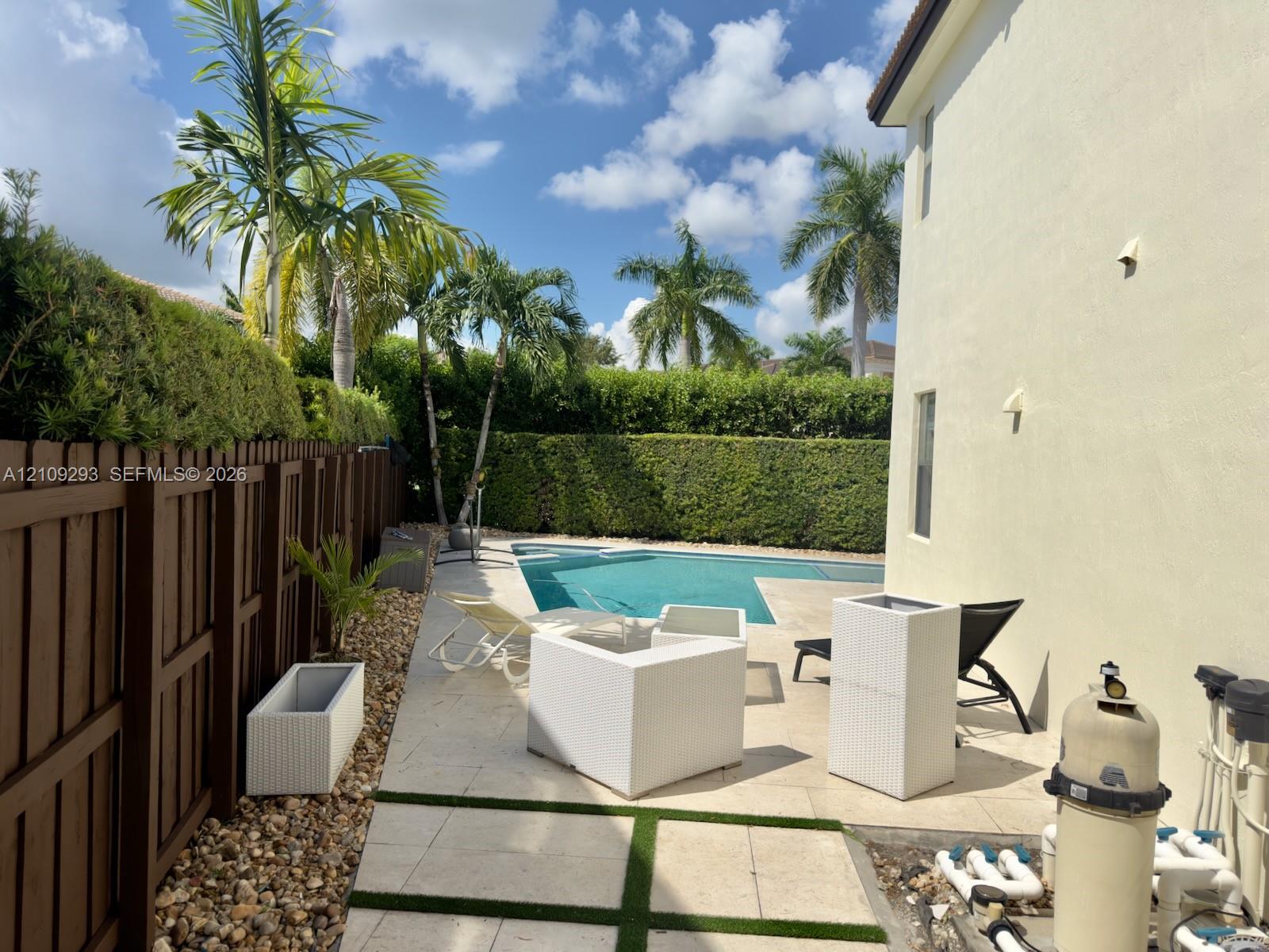 Rear yard featuring a swimming pool, stone paver decking with integrated artificial turf accents, wood-style privacy fencing, and perimeter hedge landscaping