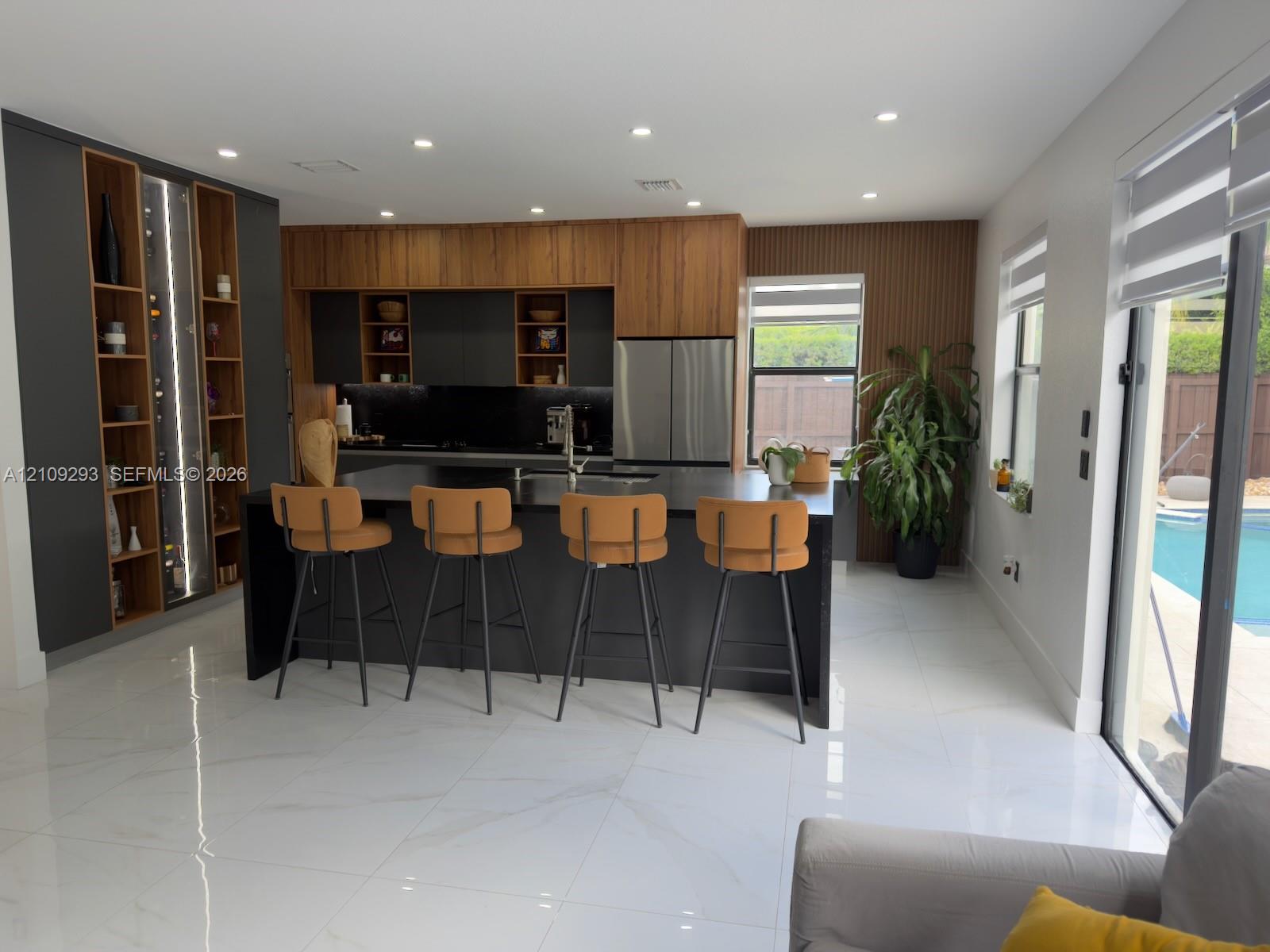 Kitchen featuring custom cabinetry with wood-finish and matte-black accents, central island with dark stone countertop, integrated floor-to-ceiling shelving, recessed lighting, and large-format tile flooring