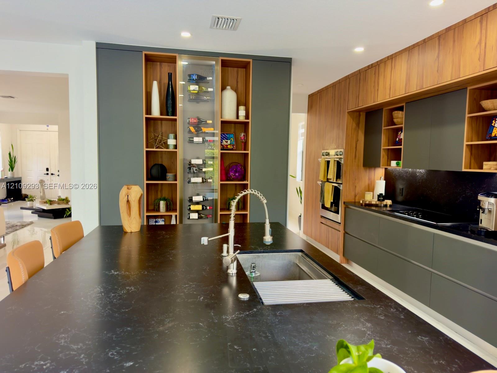 Modern kitchen featuring custom cabinetry in dark gray and wood finishes, a large island with a dark solid surface countertop, built-in stainless steel double ovens, and a dedicated vertical wine storage display