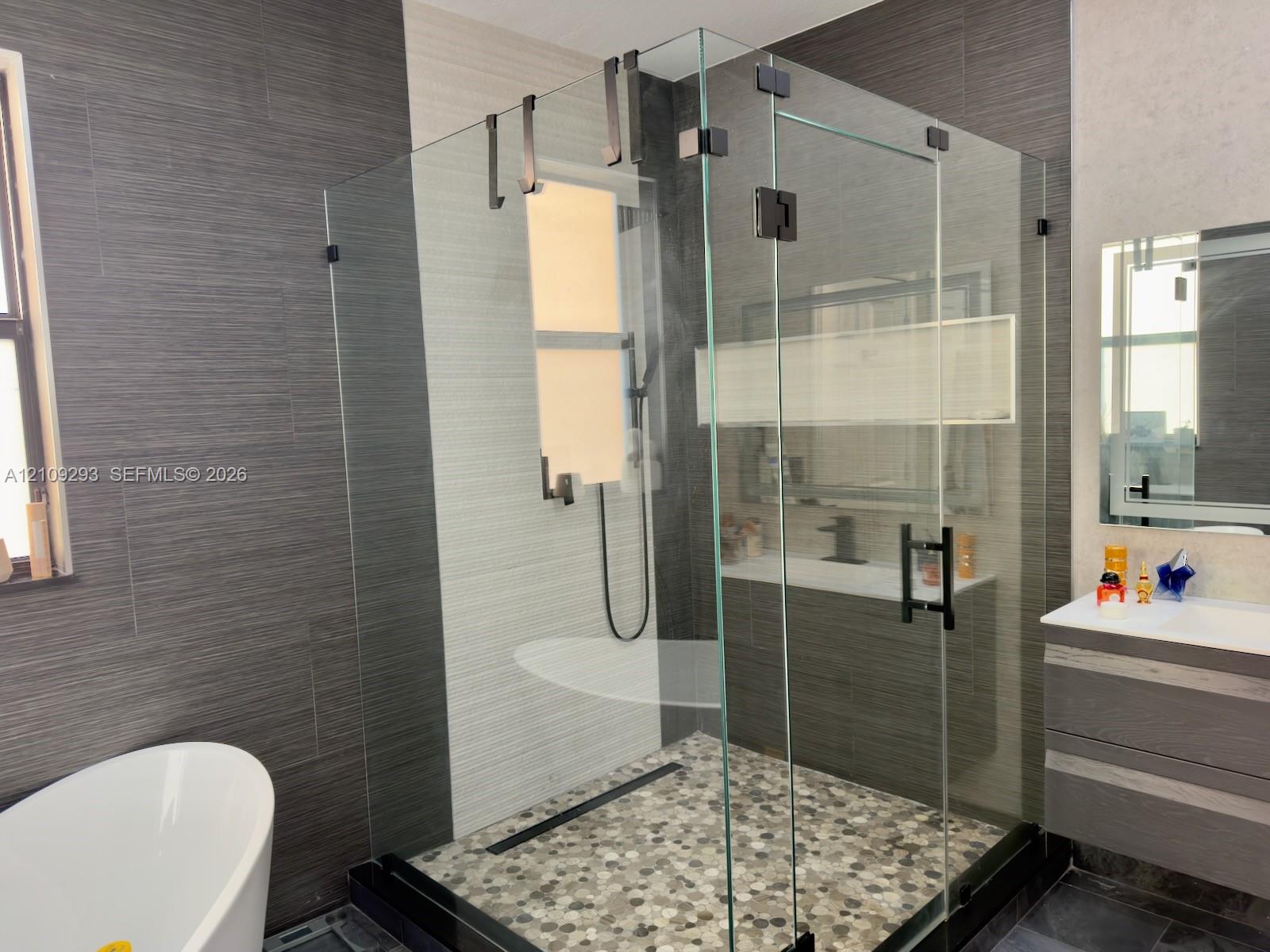 Walk-in shower featuring a frameless glass enclosure, matte black hardware, pebble-stone flooring, and a linear drain, accompanied by a floating vanity and a freestanding soaking tub