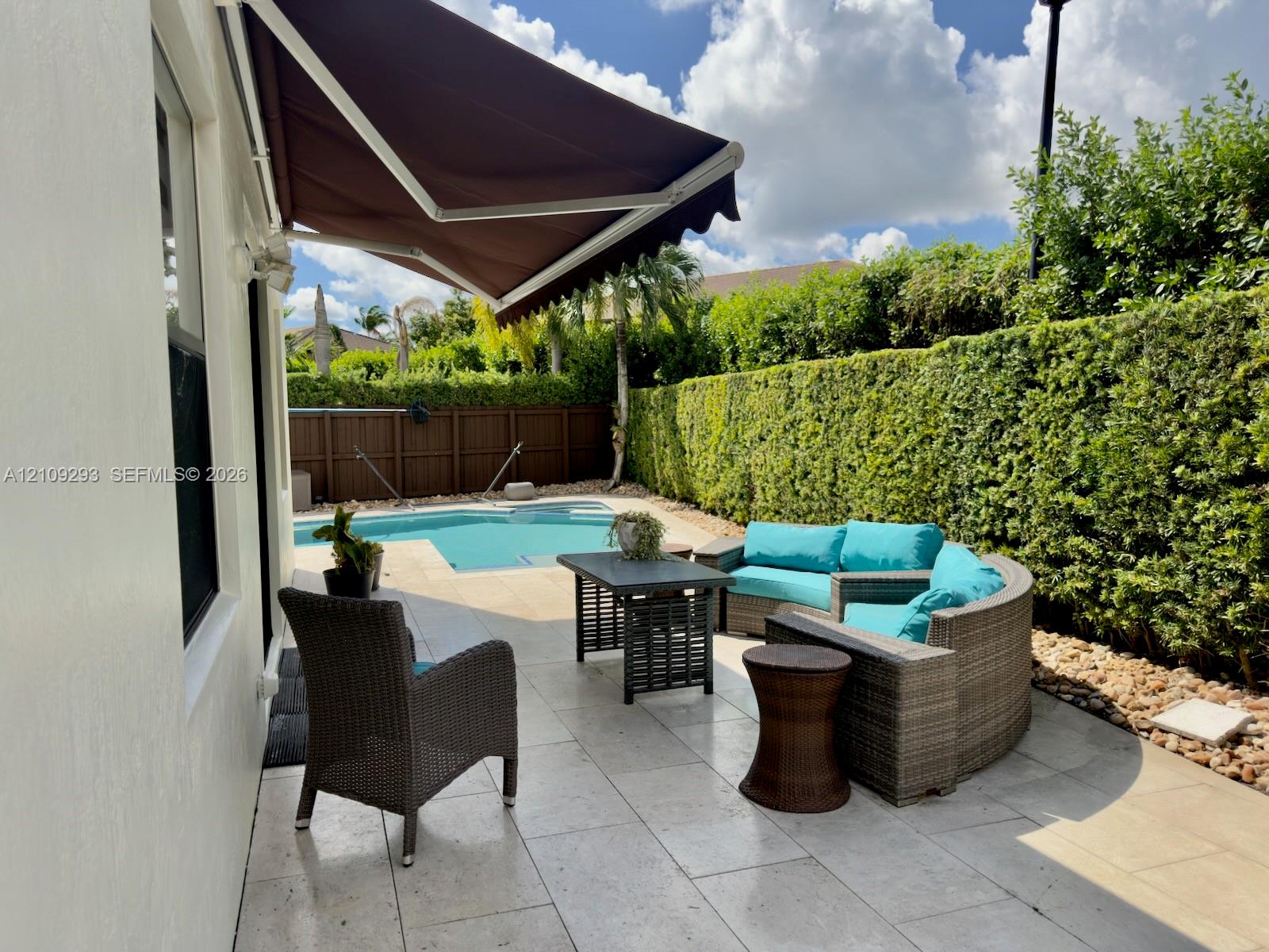 Patio space featuring light-toned stone tile flooring, a retractable fabric awning, a swimming pool, and a perimeter privacy hedge