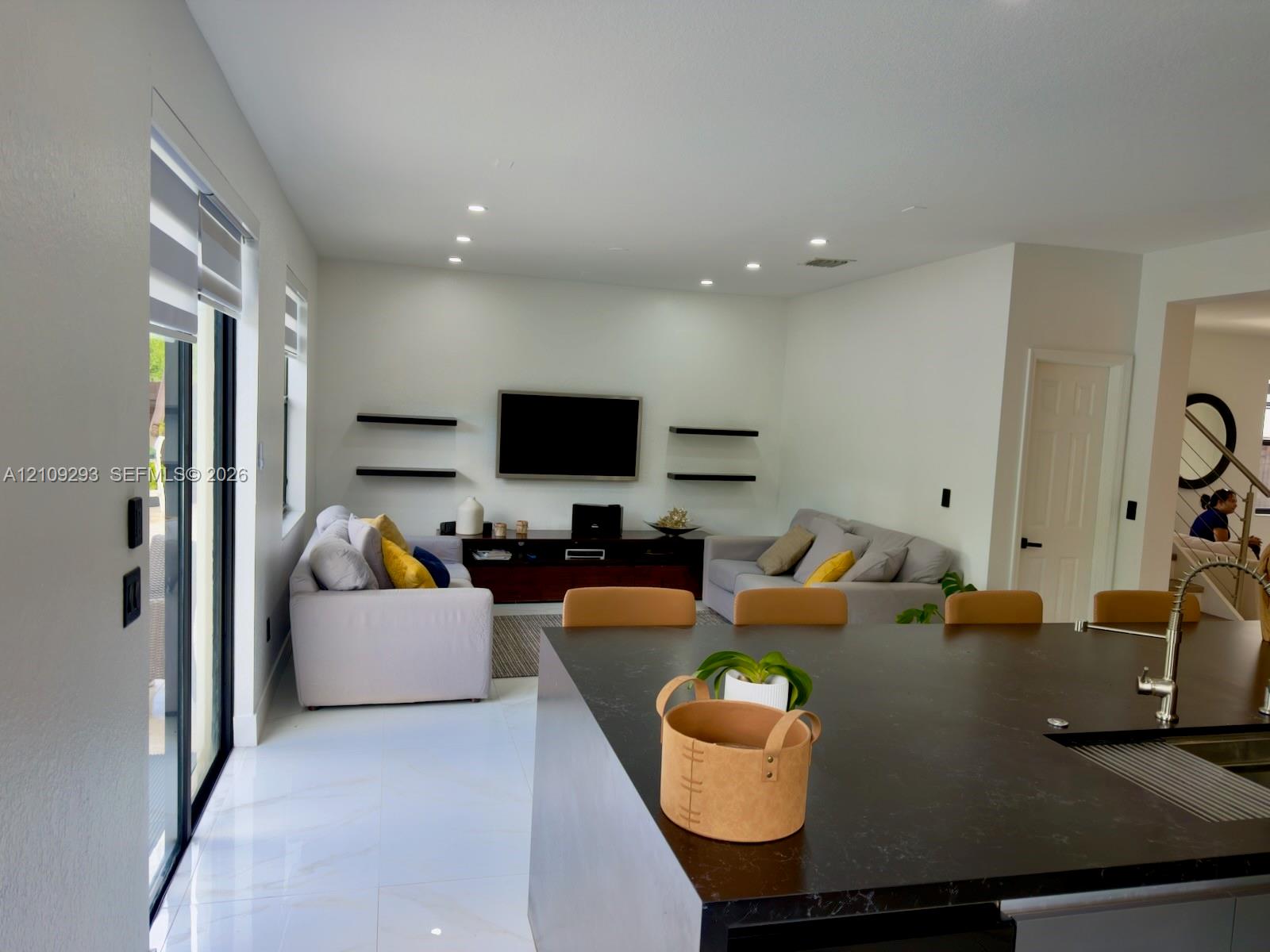 Open-concept interior featuring large-format tile flooring, recessed ceiling lighting, a dark-toned kitchen island with a metallic faucet, and matte black floating shelving