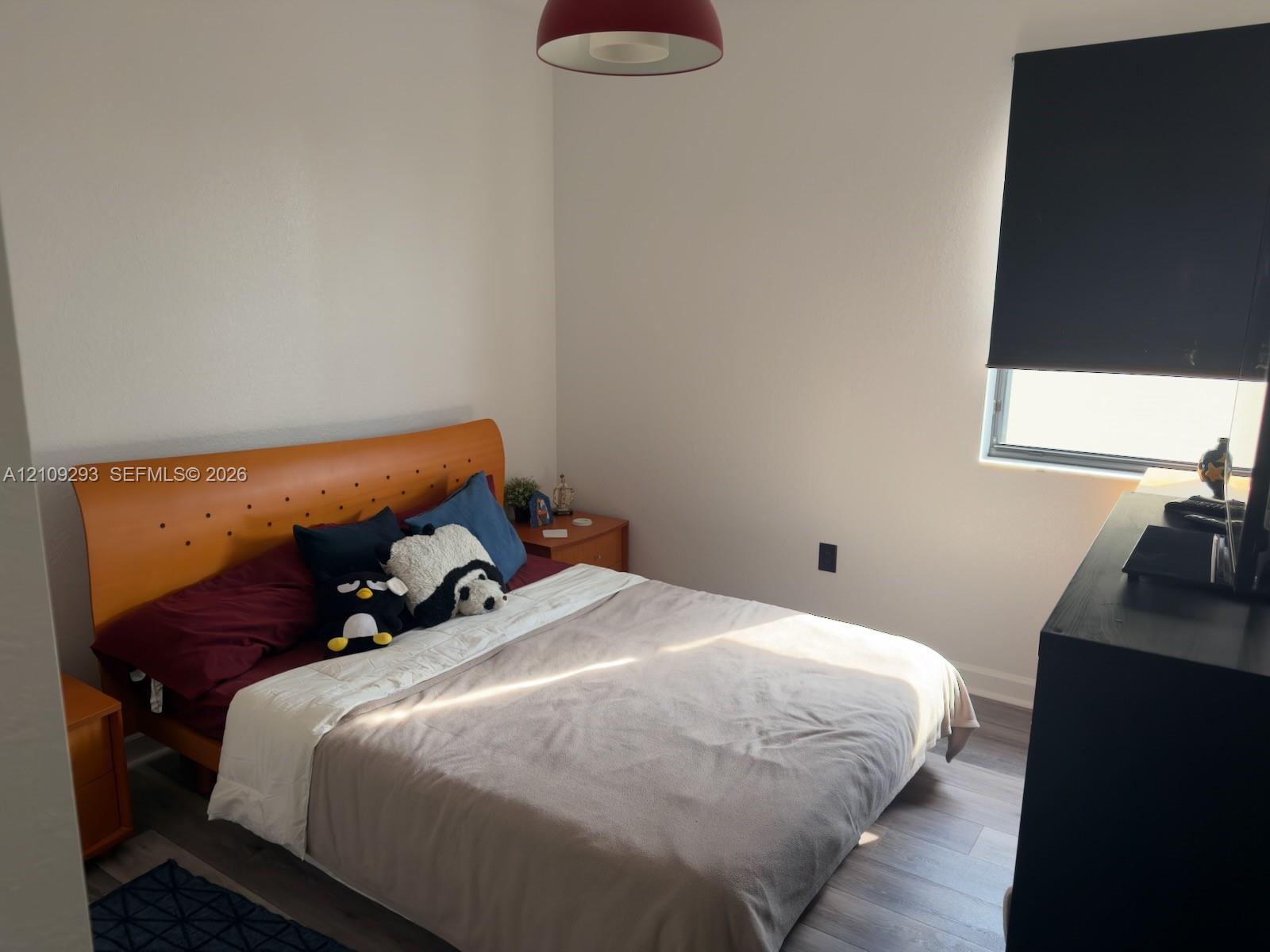 Bright bedroom featuring warm wood-finish flooring, an upholstered platform bed with button-tufted headboard, and ample natural light from a window with blackout shade